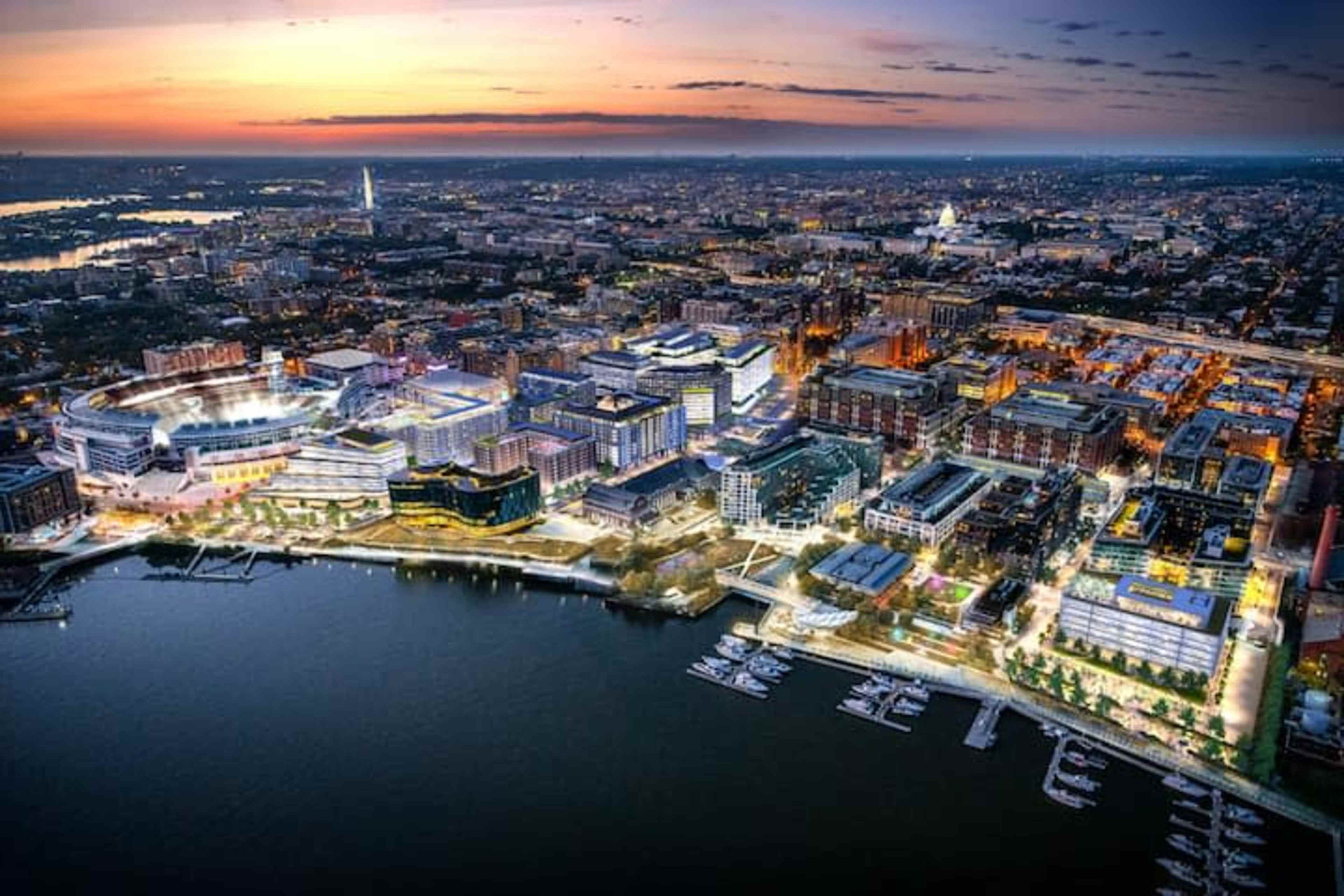 An aerial view captures a bustling waterfront city at sunset, showcasing illuminated buildings, a marina, and a vibrant skyline.