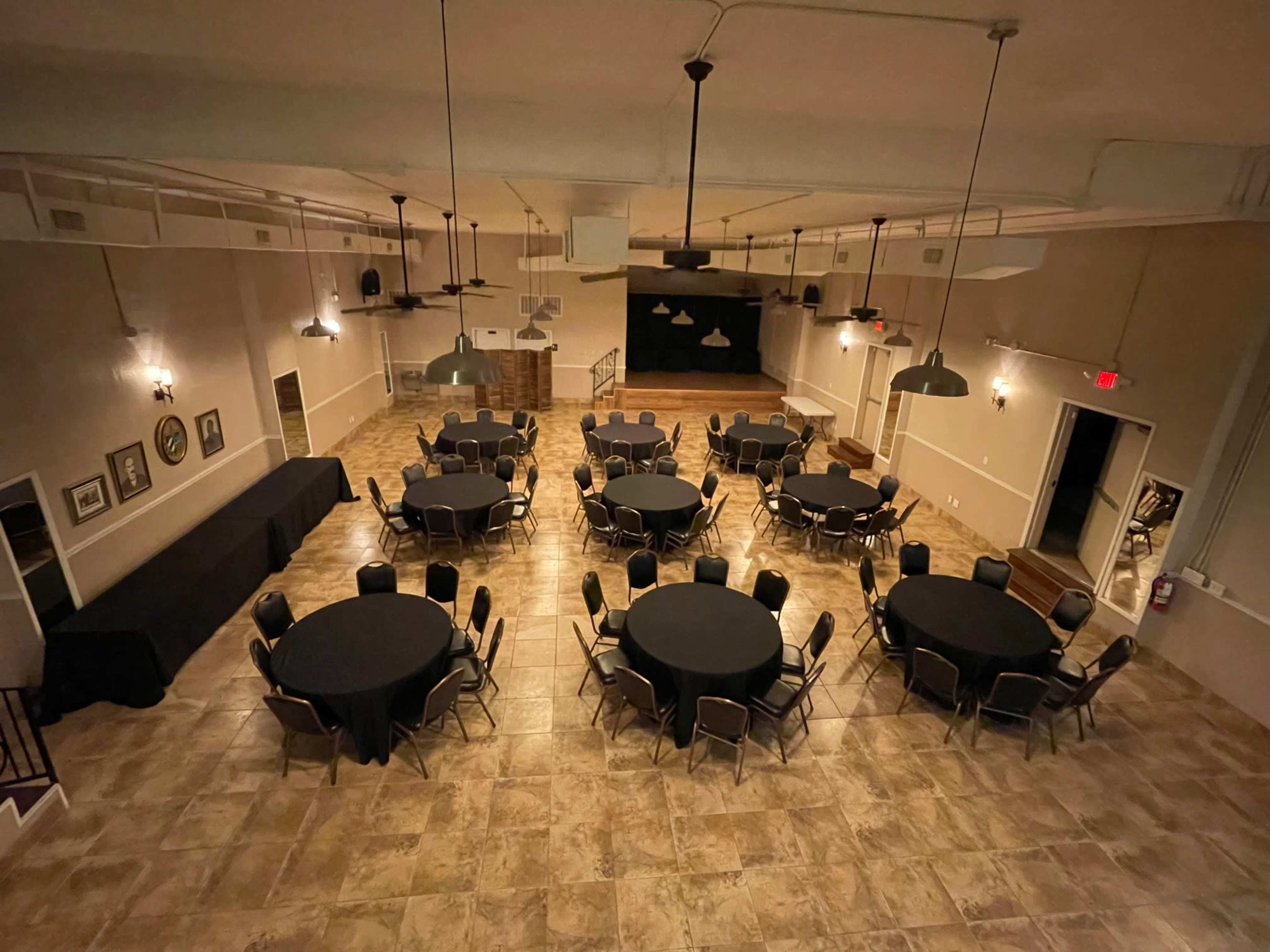 A spacious event room set up with multiple round tables covered in black tablecloths, arranged in a grid pattern on a tiled floor.