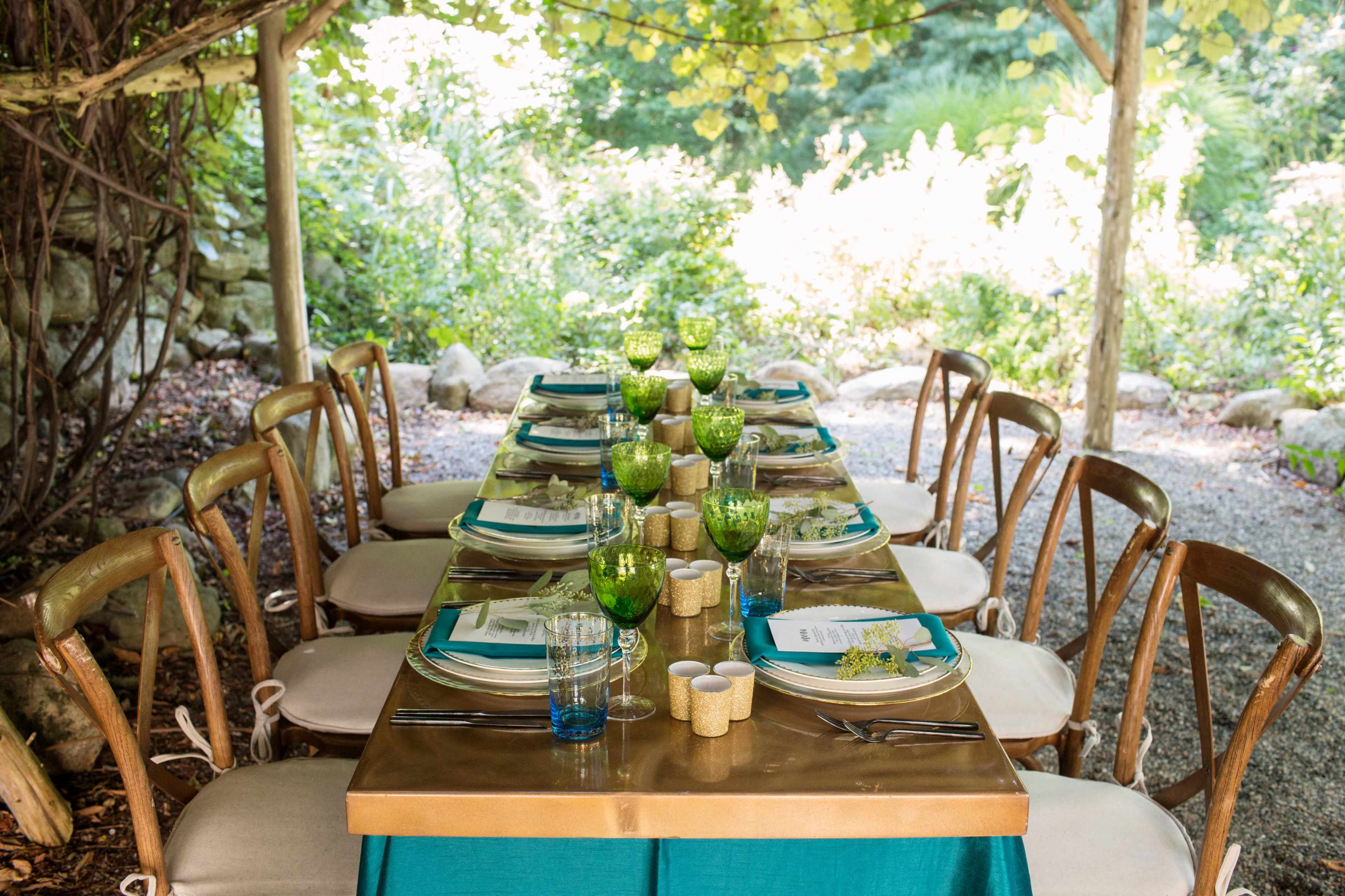 A long dining table is set outdoors under a canopy, adorned with green glassware, plates, and floral centerpieces amidst a natural garden backdrop.