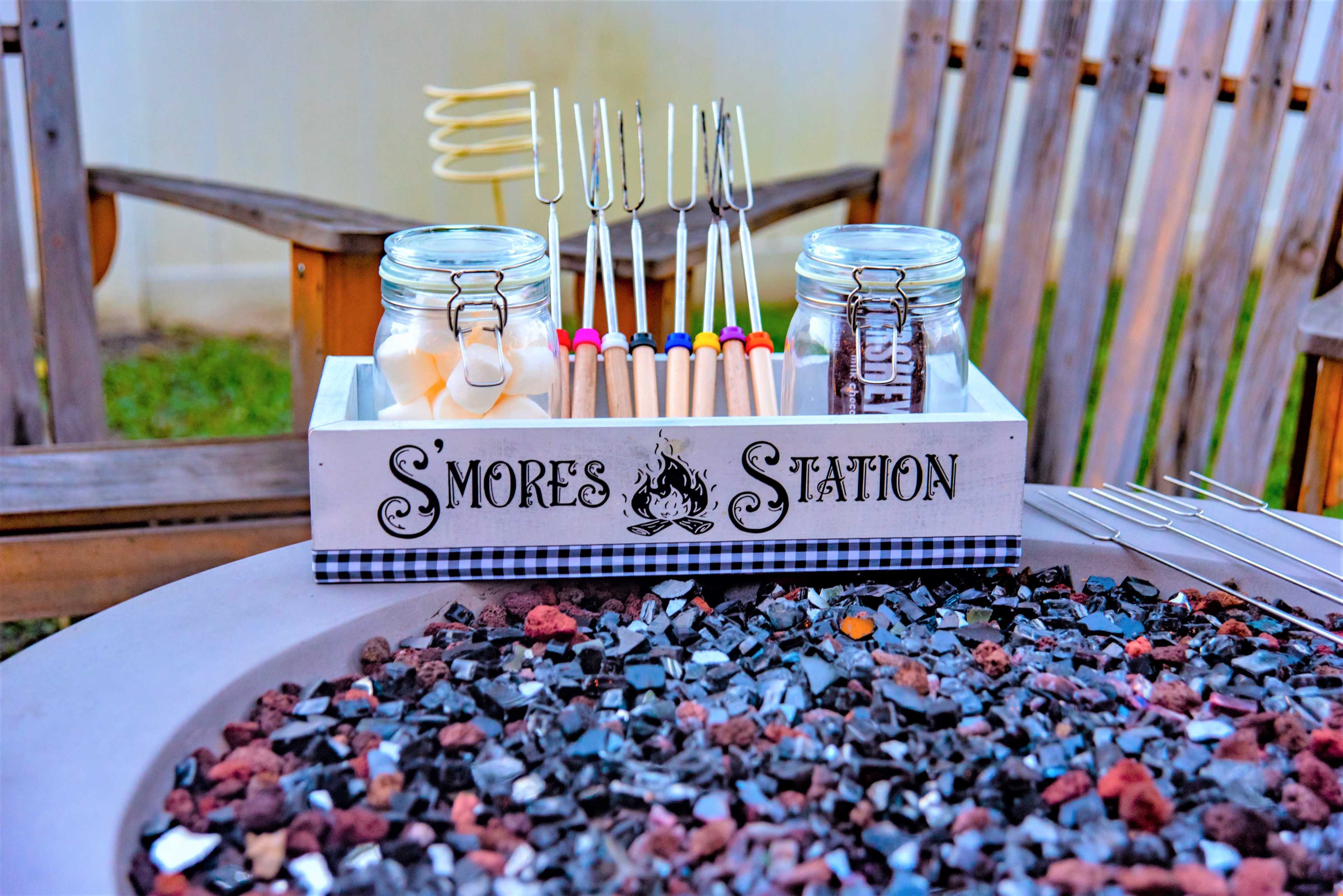 A S'mores station featuring jars of graham crackers, marshmallows, and chocolate, along with skewers, is set up on a fire pit table.