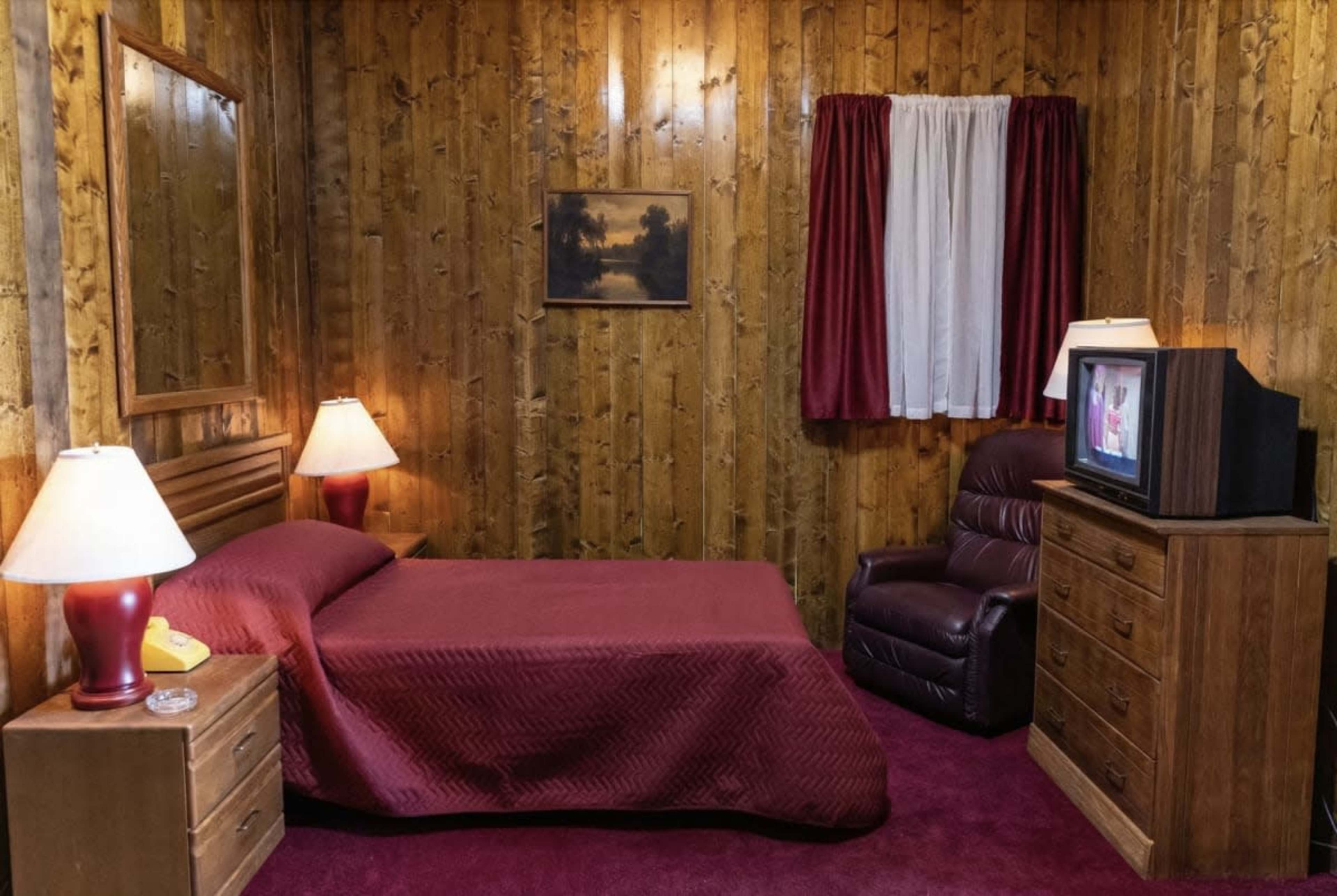 The image shows a small bedroom with wooden paneling, a red bedspread, a recliner chair, a television, and a nightstand with lamps.