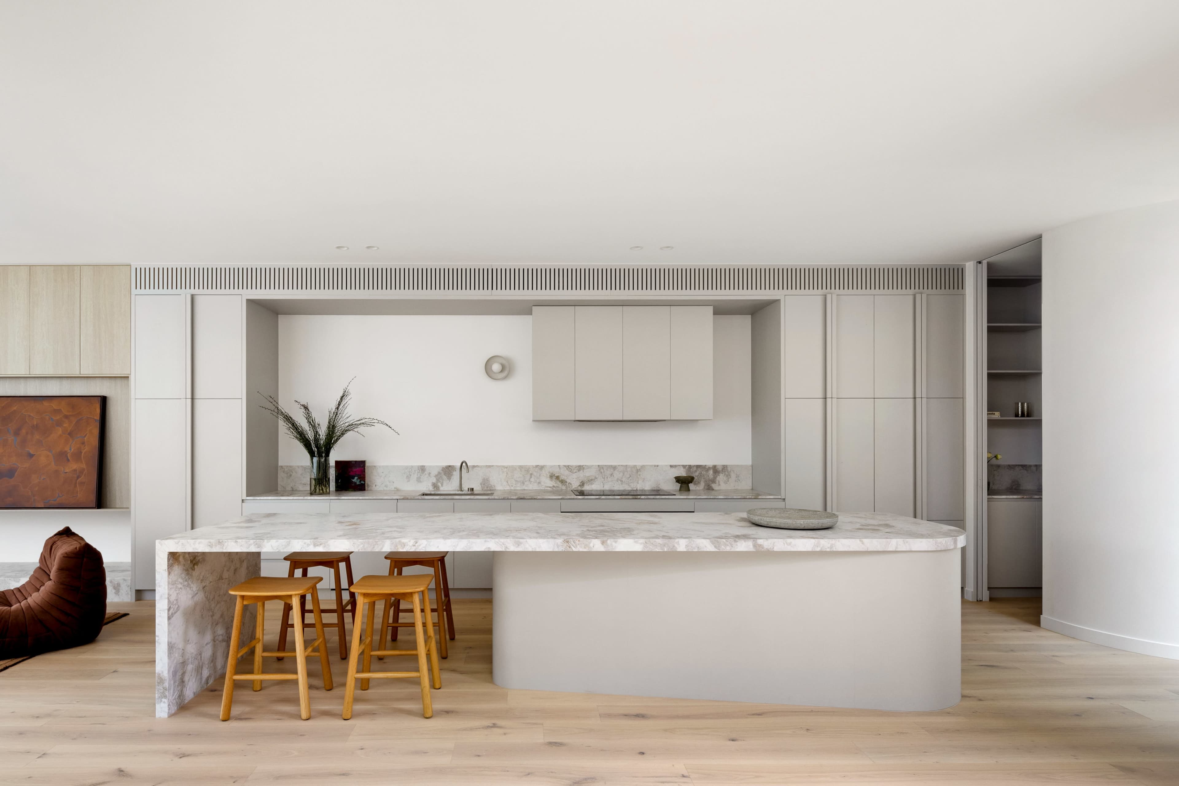 The image shows a modern kitchen with a large marble island, wooden bar stools, and minimalist cabinetry.