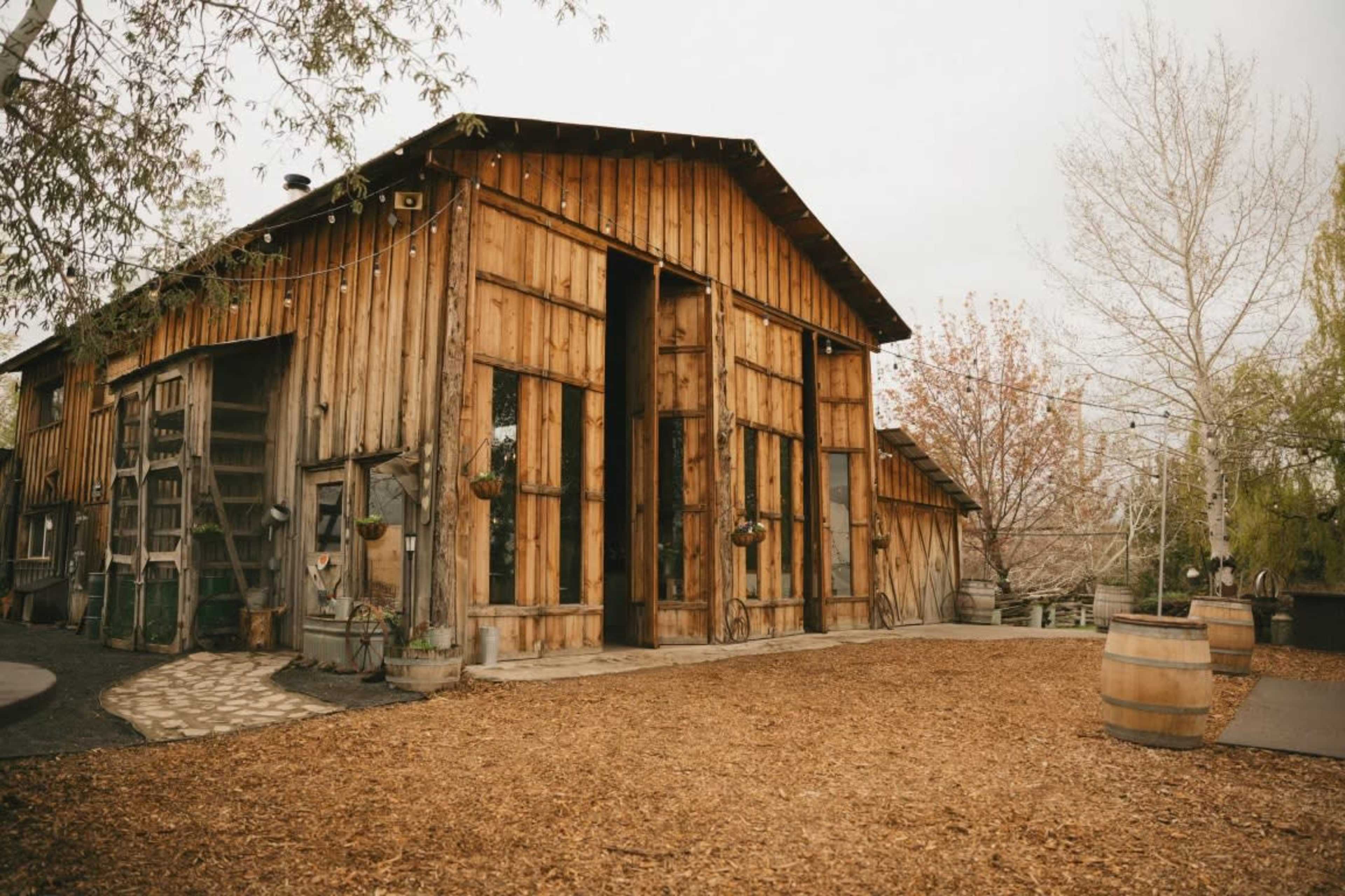 The image shows a wooden barn with large open doors, surrounded by mulch and trees.