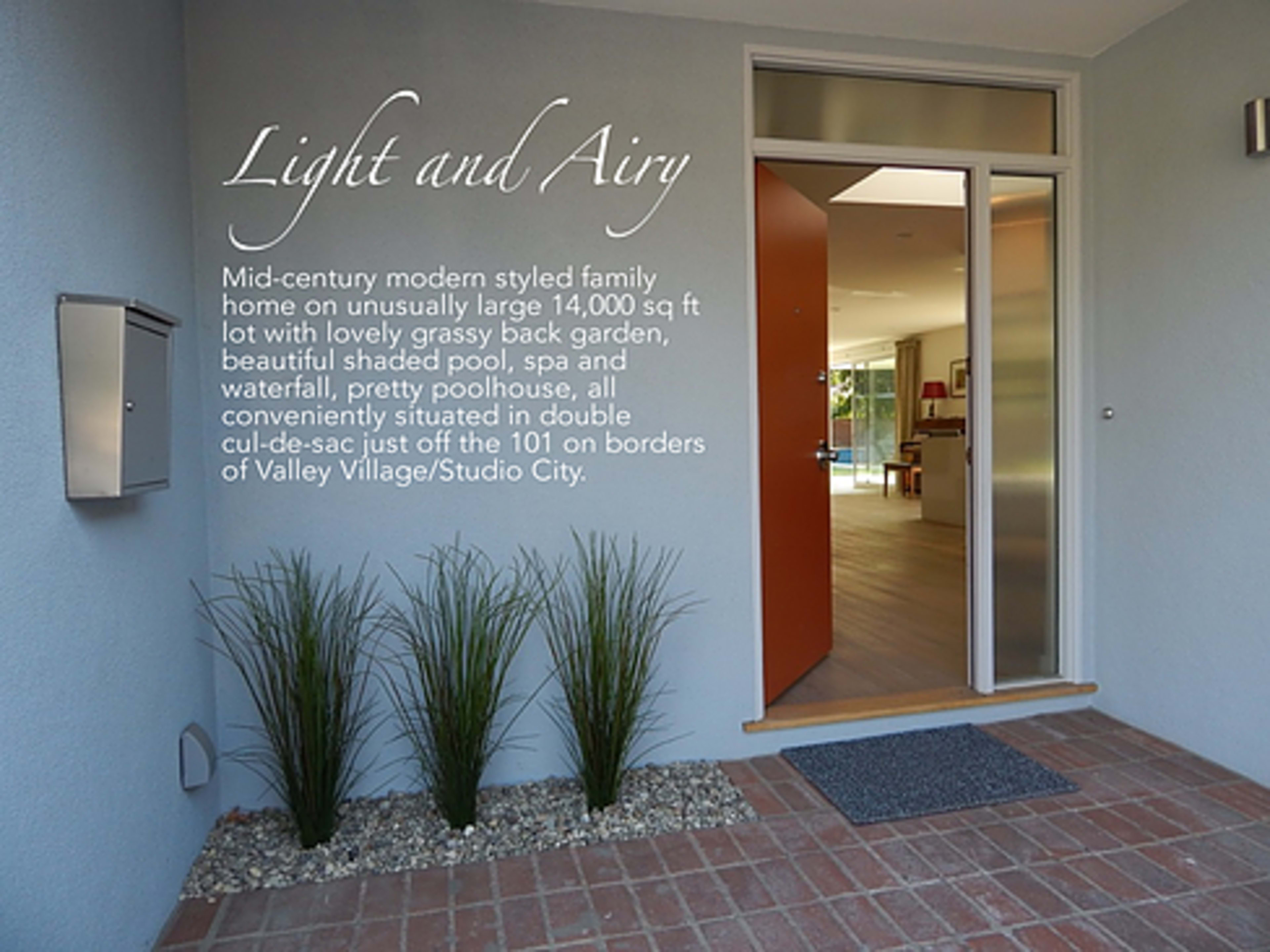 The entrance of a mid-century modern styled family home, featuring an open door, a pathway, and decorative plants alongside a wall with a descriptive sign.