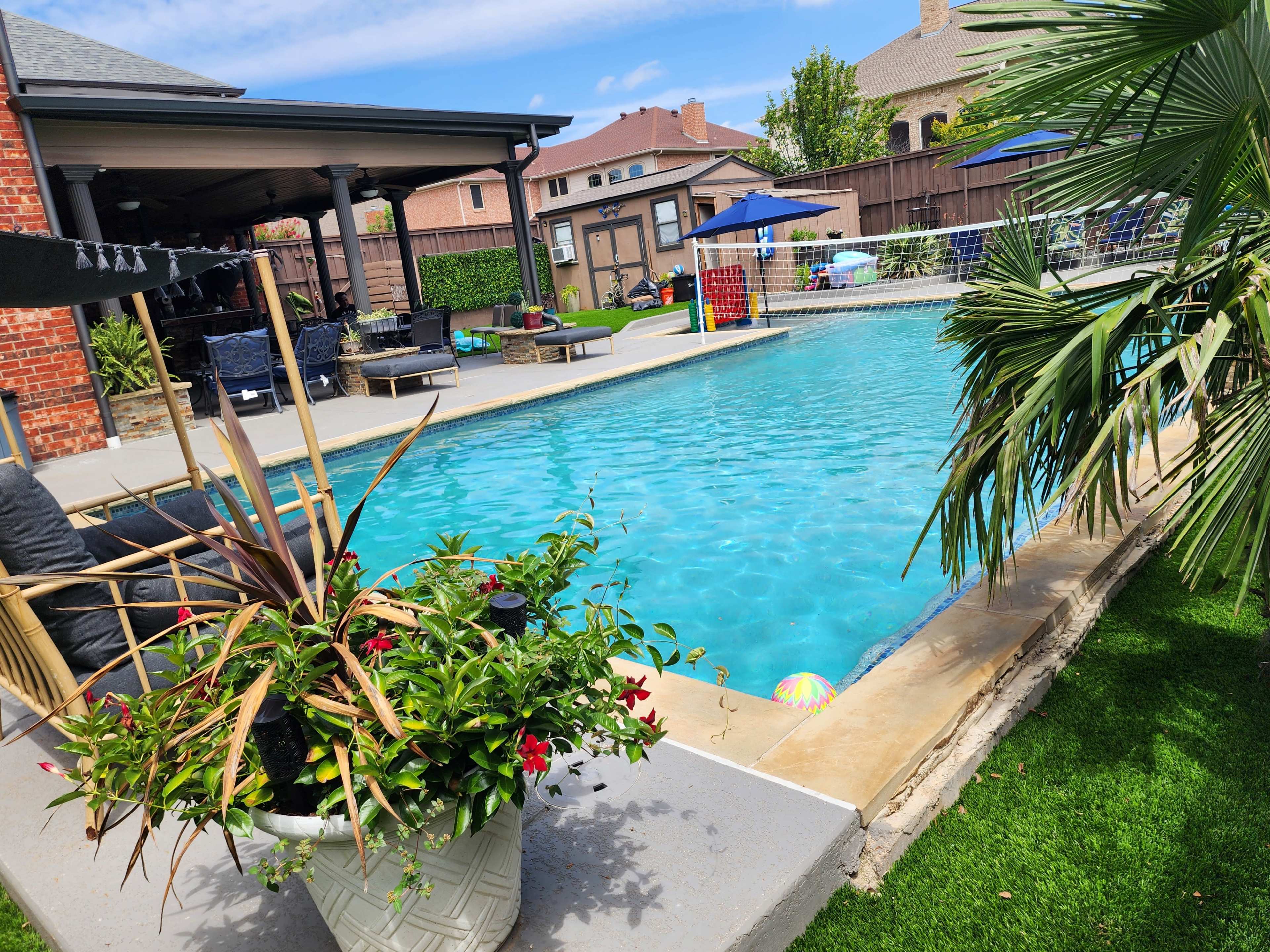 The image shows a backyard swimming pool surrounded by lounge chairs, umbrellas, and a well-maintained lawn with potted plants.