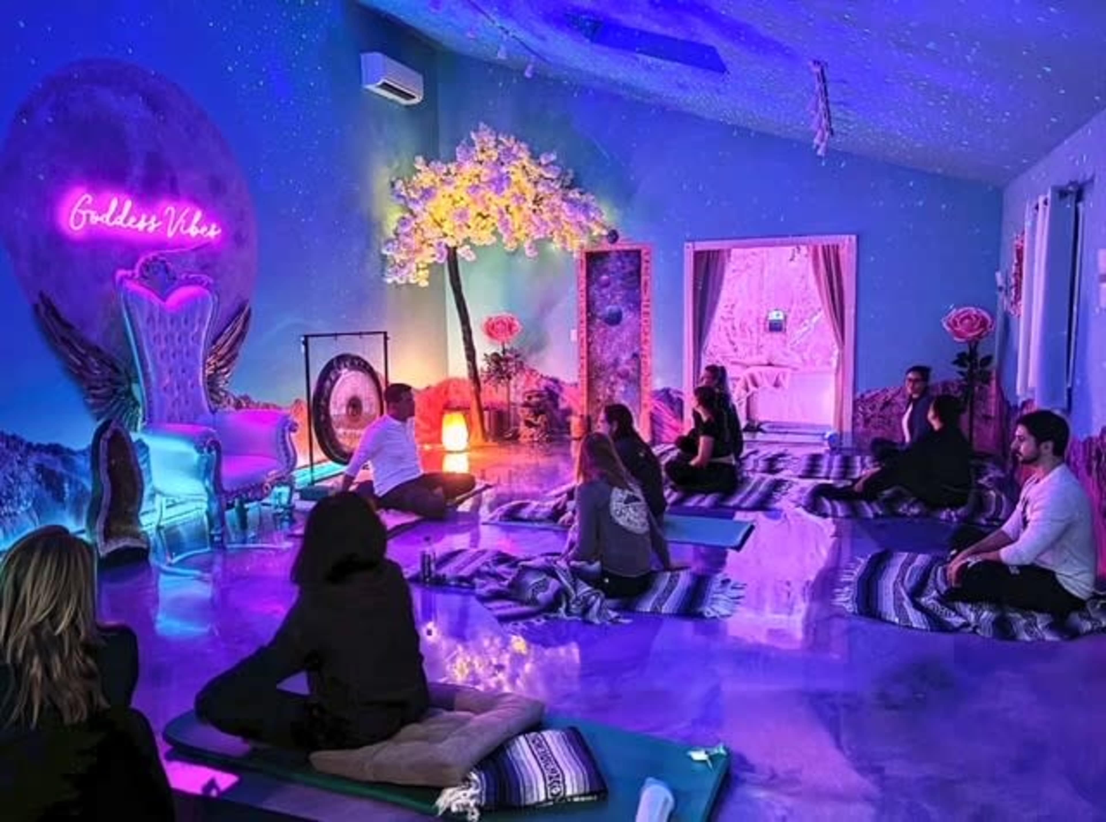 A group of people sits on mats in a brightly colored room adorned with neon lights, flowers, and a decorative arch.