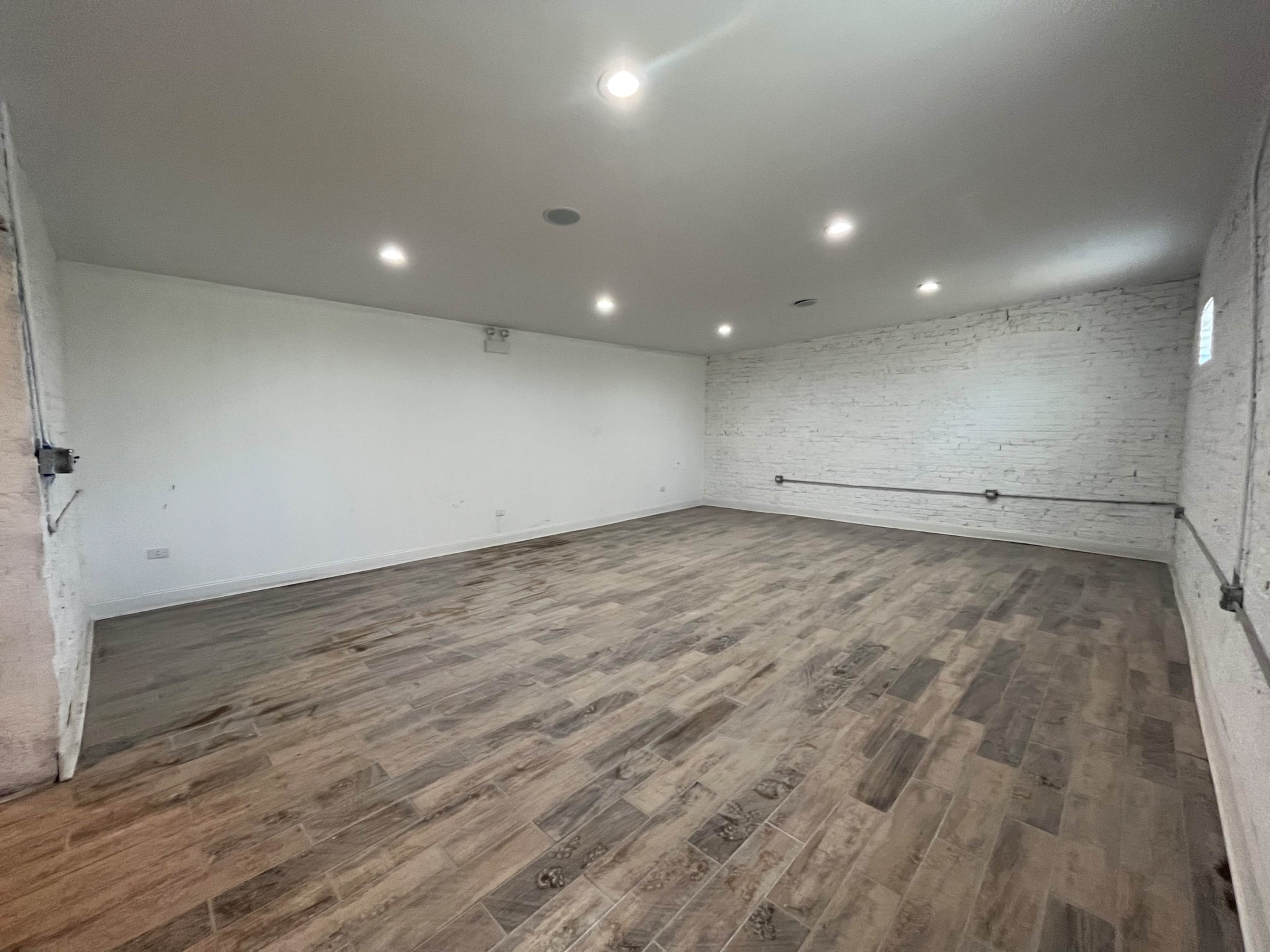 An empty room with white brick walls and wooden flooring, illuminated by several ceiling lights.