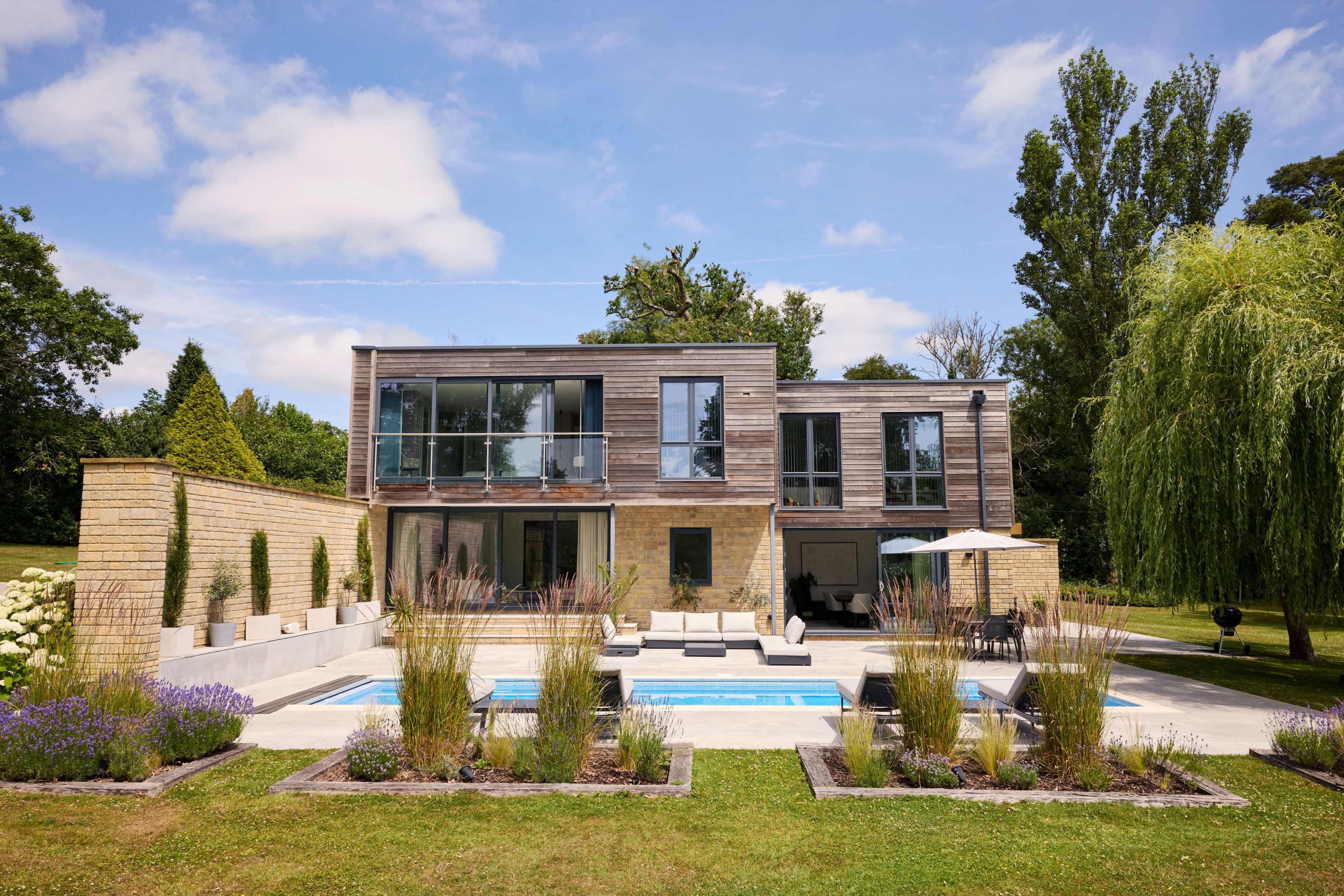 The image shows a modern two-story house with large windows, surrounded by a landscaped garden featuring a swimming pool and various plants.