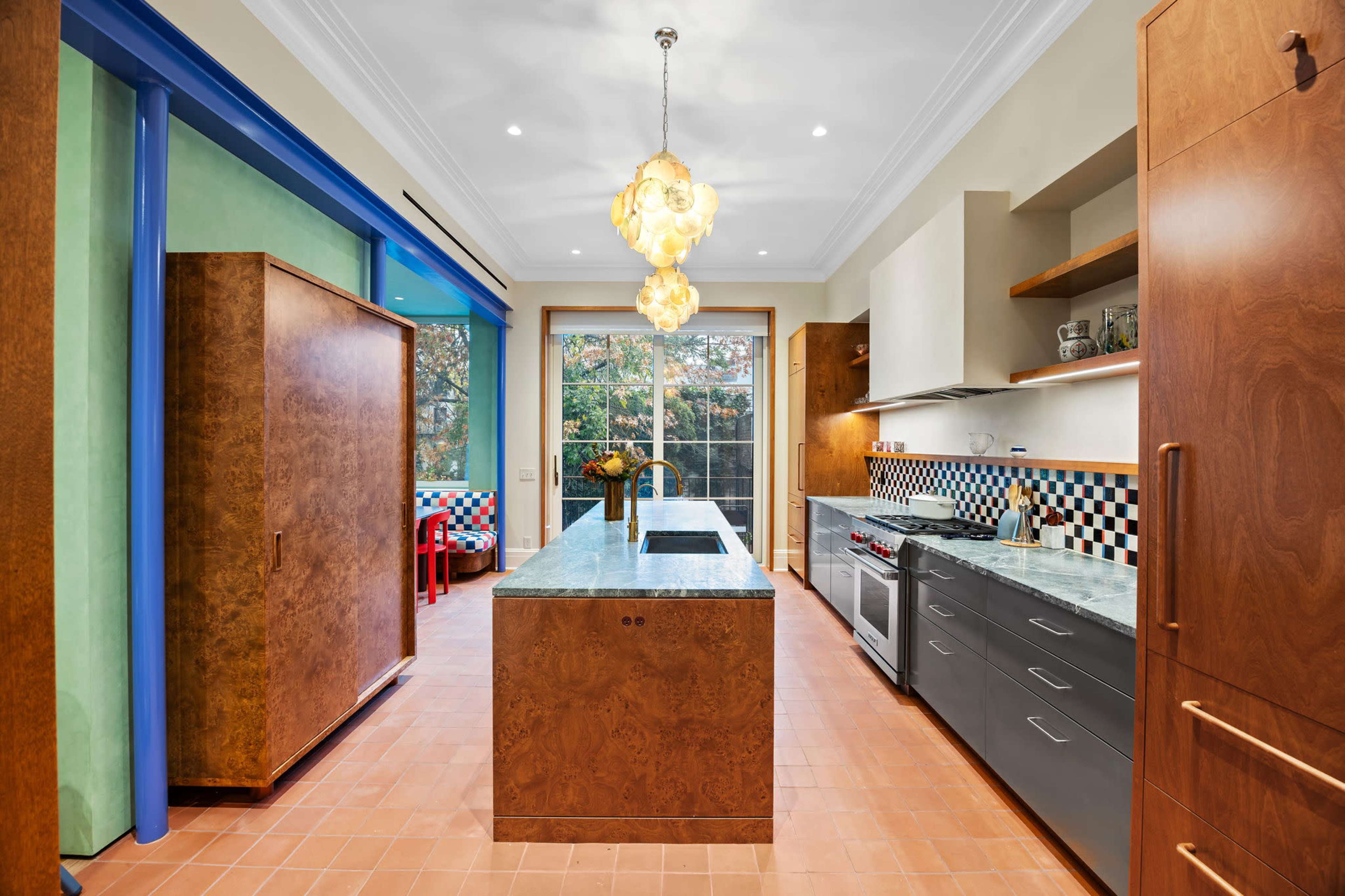 The modern kitchen features a large island with a stone countertop, wooden cabinetry, and colorful decorative elements, with large windows allowing natural light to fill the space.
