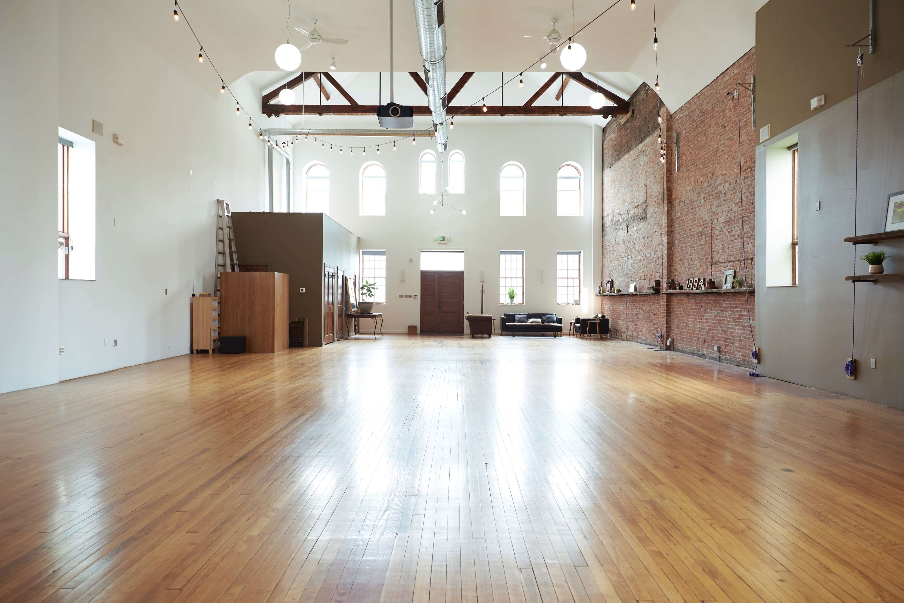 A spacious, wooden-floored room with high ceilings, large windows, and a mix of brick and smooth walls.
