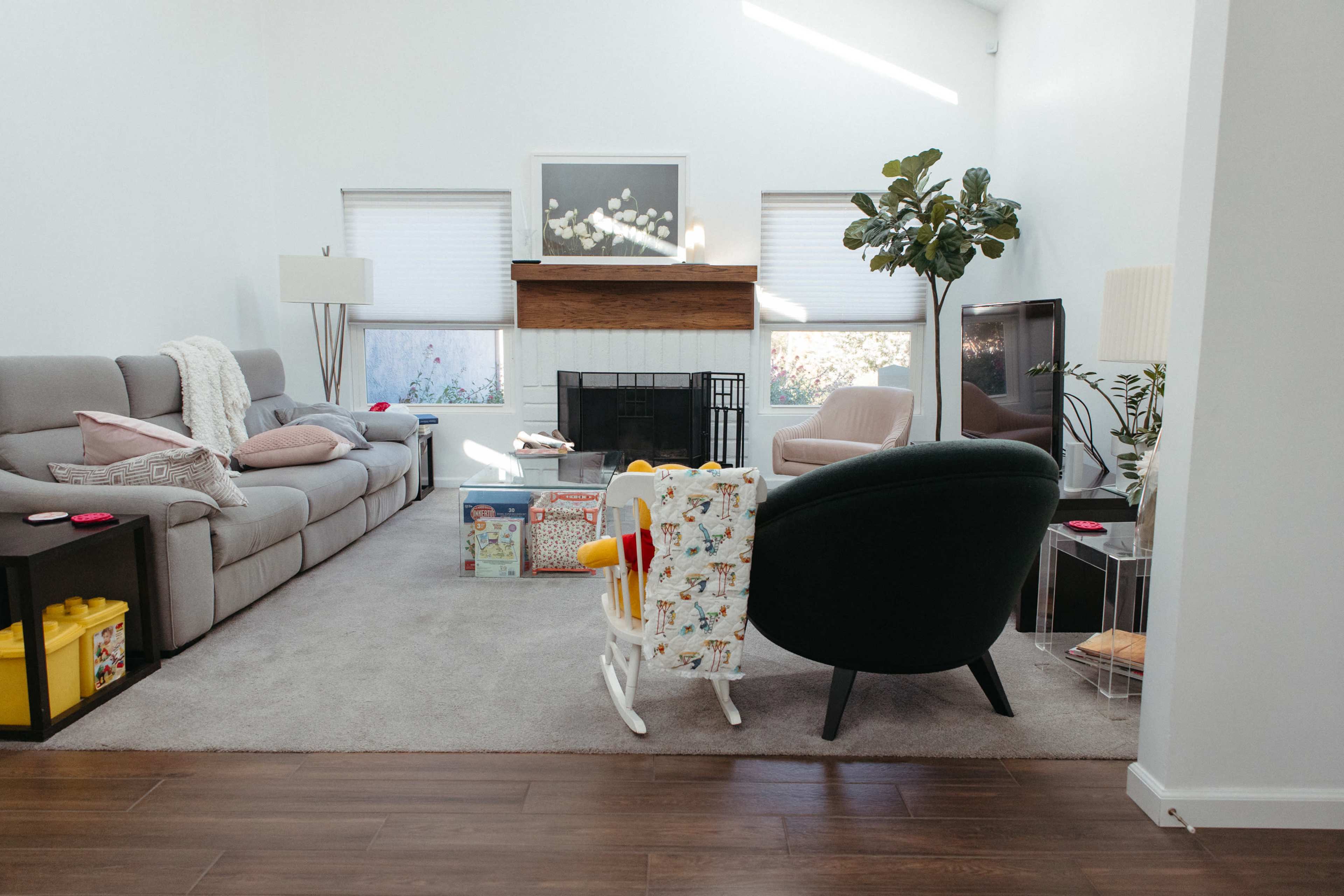 The image shows a modern living room with a gray sofa, a pink chair, a fireplace with a wooden mantel, and a variety of decorative items, including plants and toys.