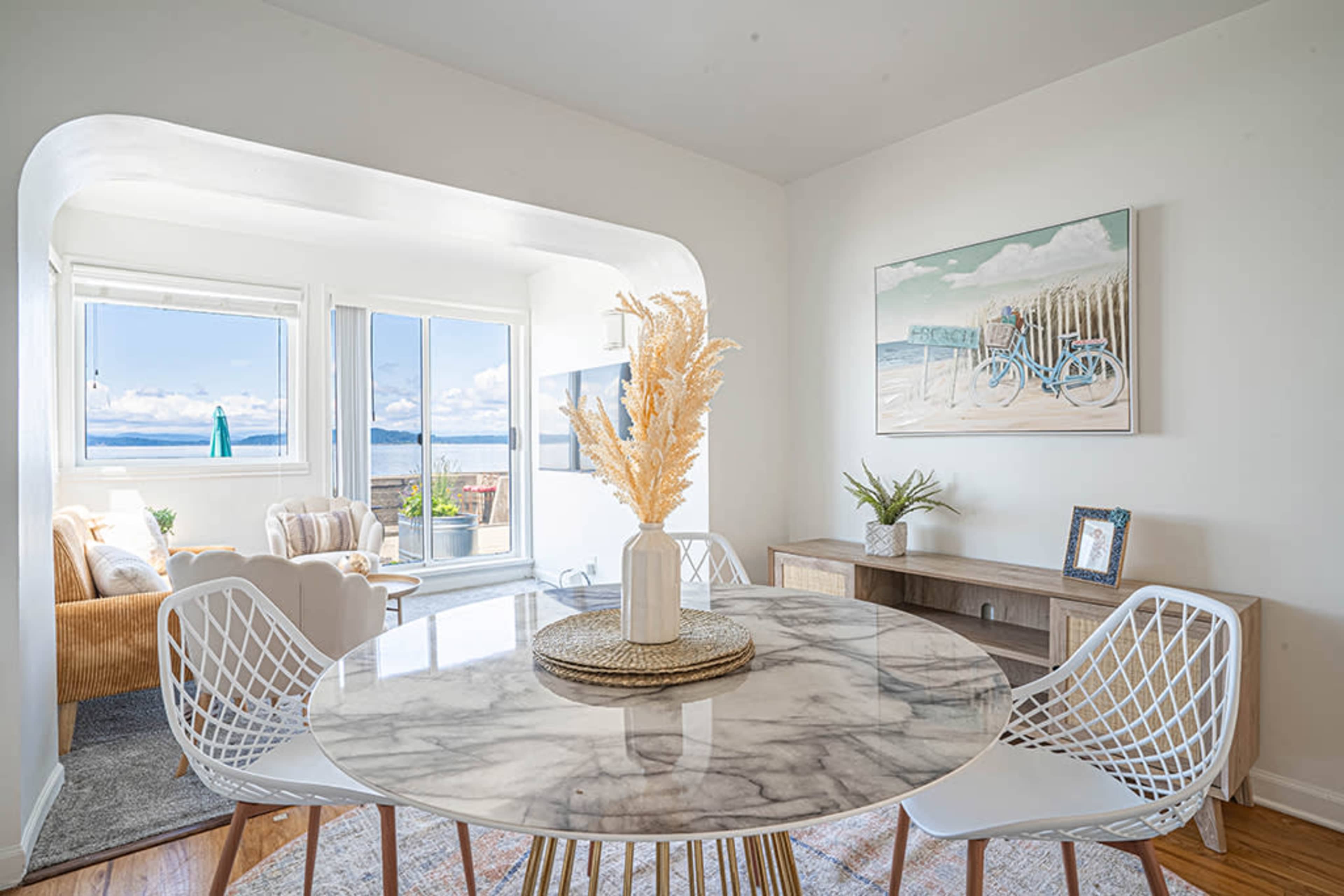 A modern dining area features a round marble table with white chairs, complemented by a decorative vase and a view of the ocean through large windows.