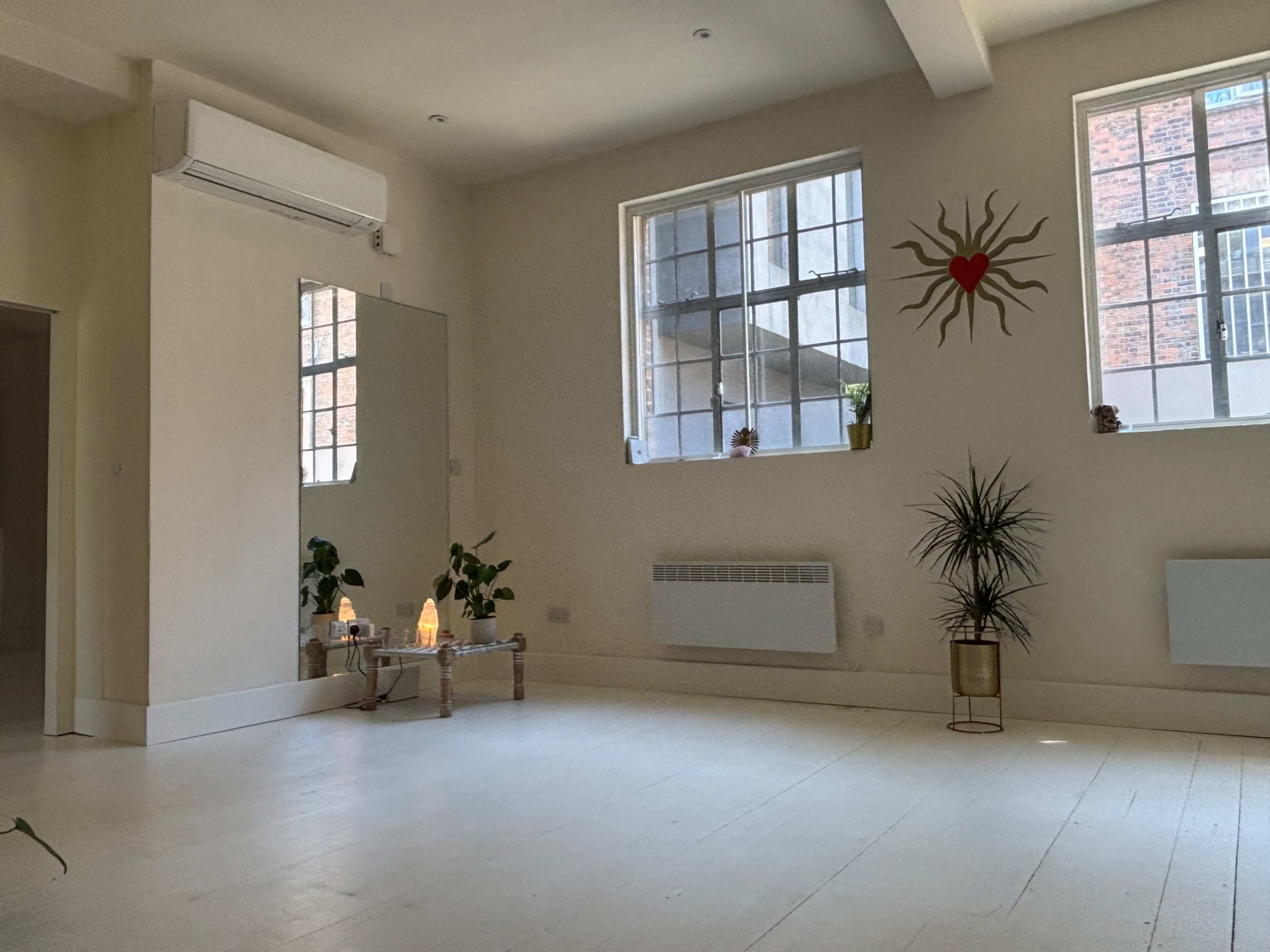 The image shows a spacious, warmly lit room with large windows, a mirror, plants, and decorative elements including a heart inside a sun design on the wall.