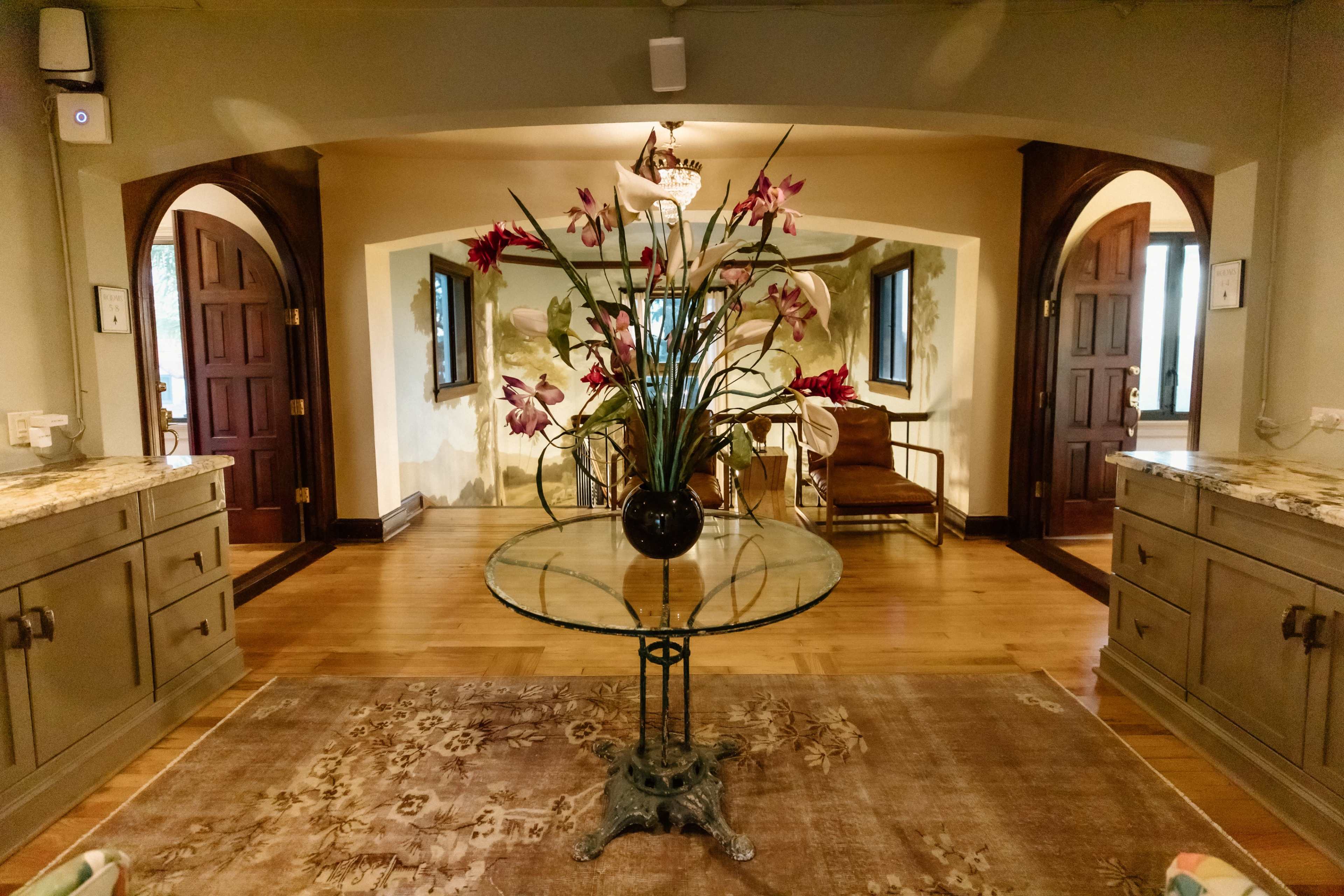 A glass table with a floral arrangement stands in the center of a room with arched doorways and wooden furniture.