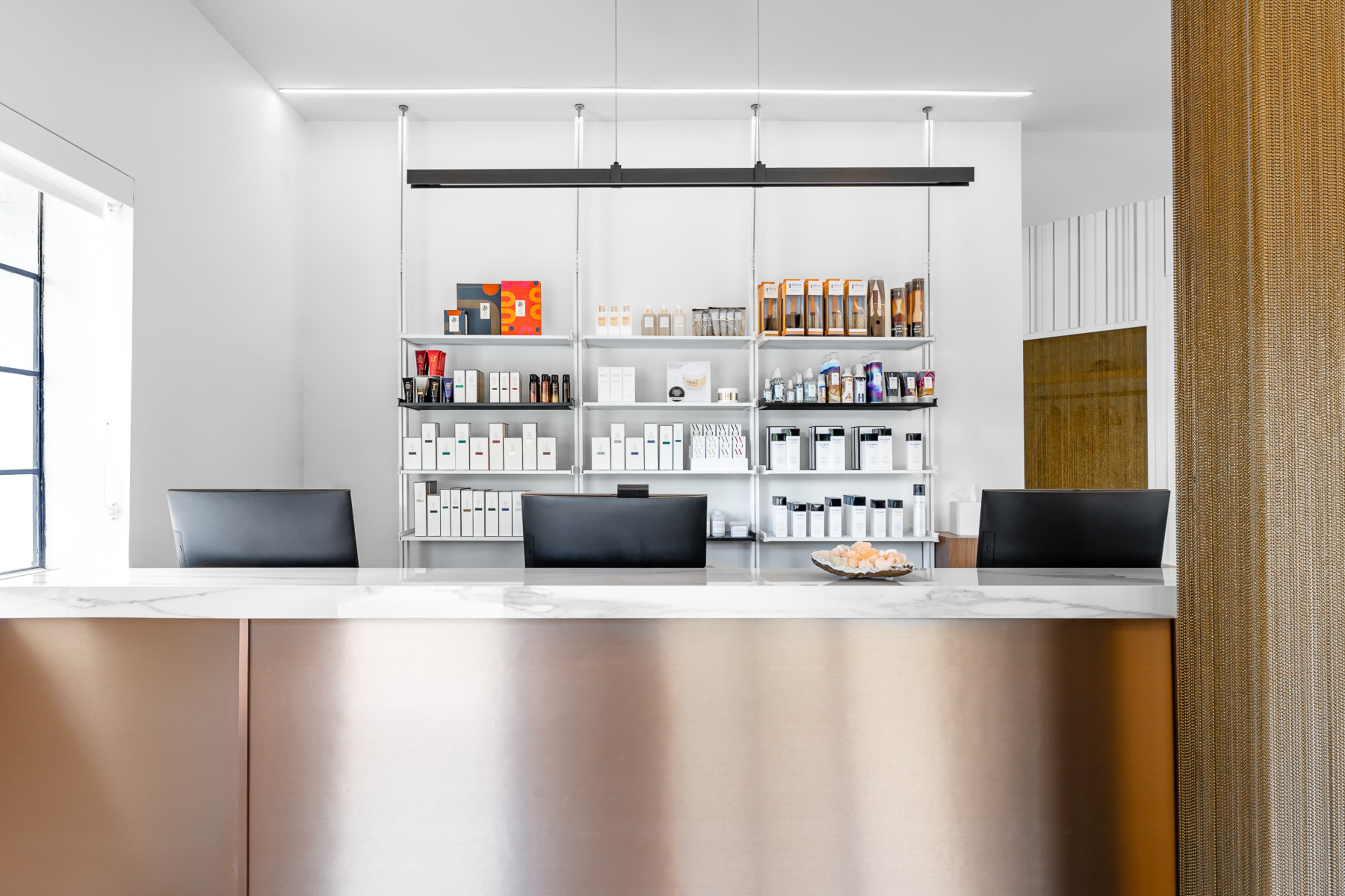 A modern reception desk with three black chairs and a display of neatly arranged skincare products on shelves in the background.
