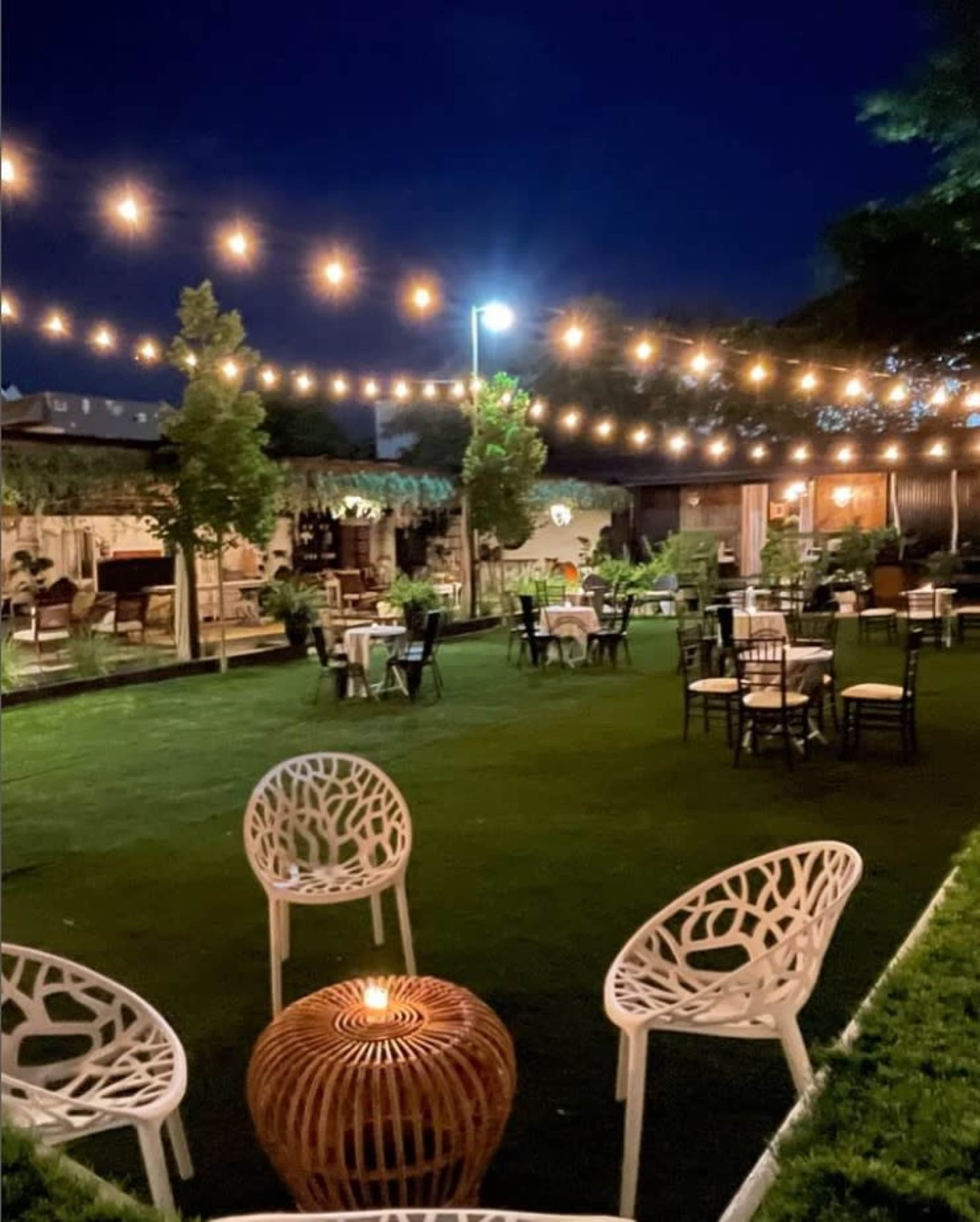 An outdoor restaurant with string lights overhead, featuring green grass, circular tables, and white chairs arranged around a central fire pit.