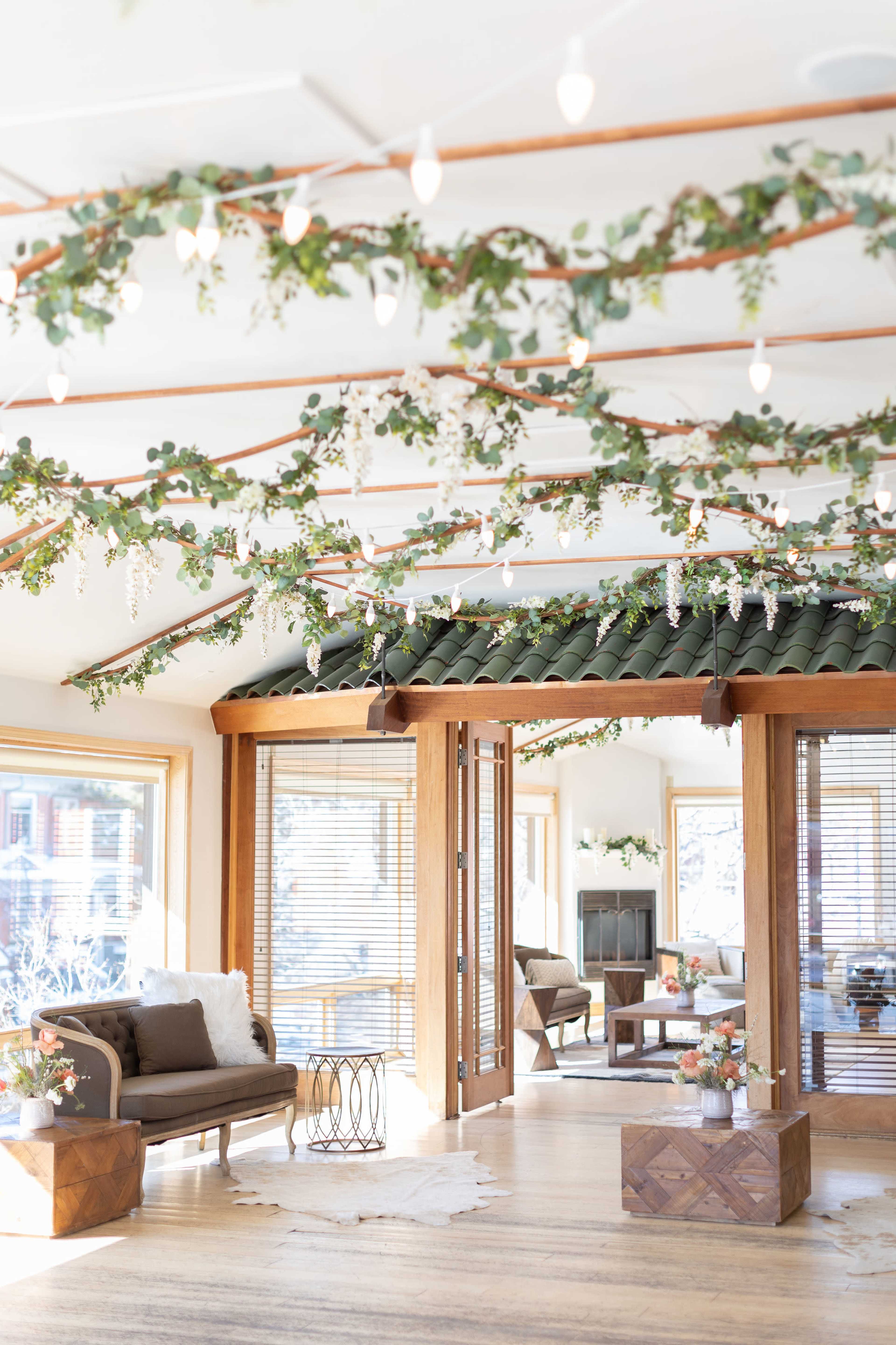 A cozy room features a wooden ceiling adorned with greenery and string lights, with furniture arranged around a central area near large windows.
