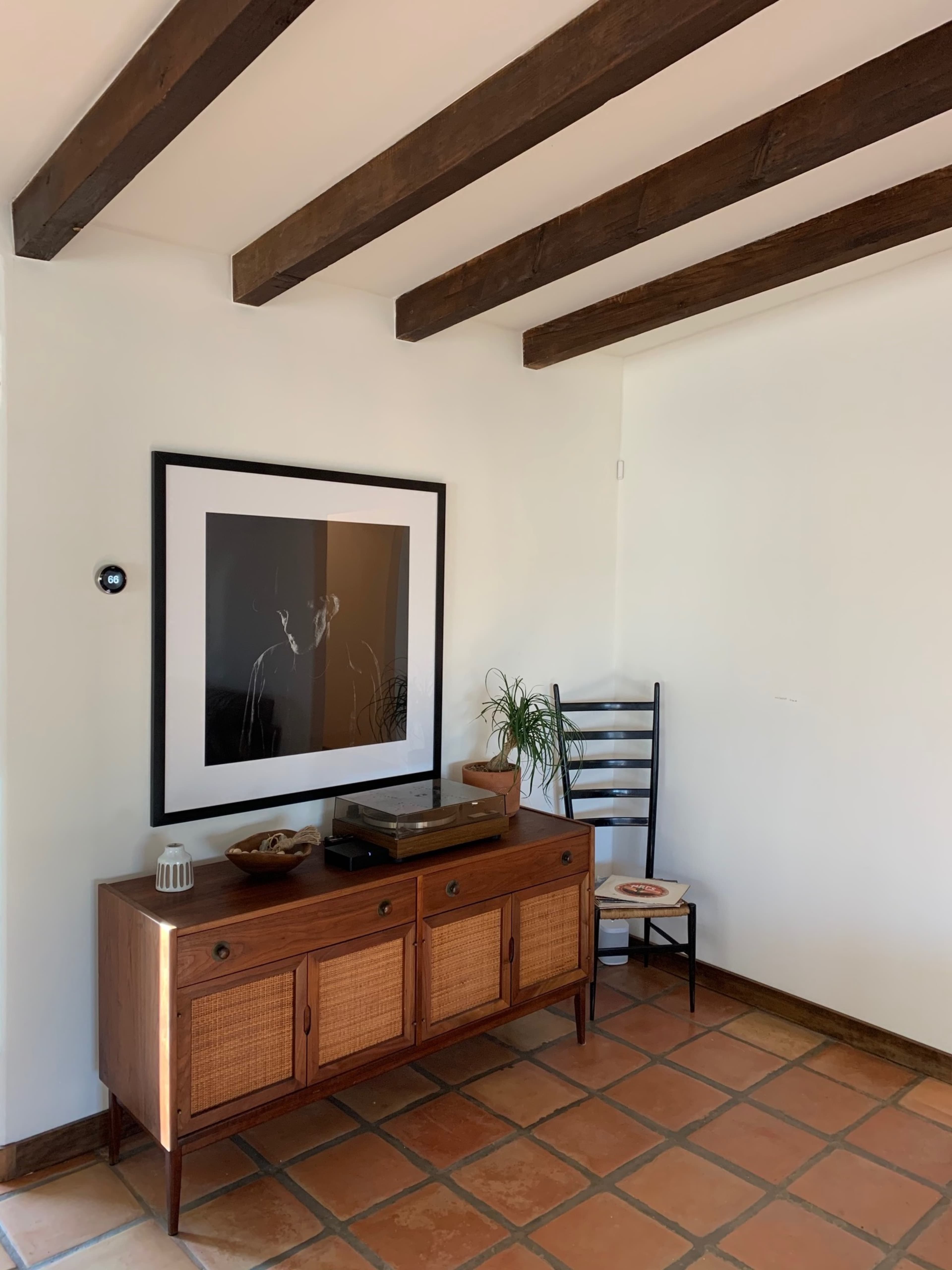 A corner of a room features a wooden sideboard with a record player, a framed black-and-white photograph on the wall, a simple chair, and a small potted plant.