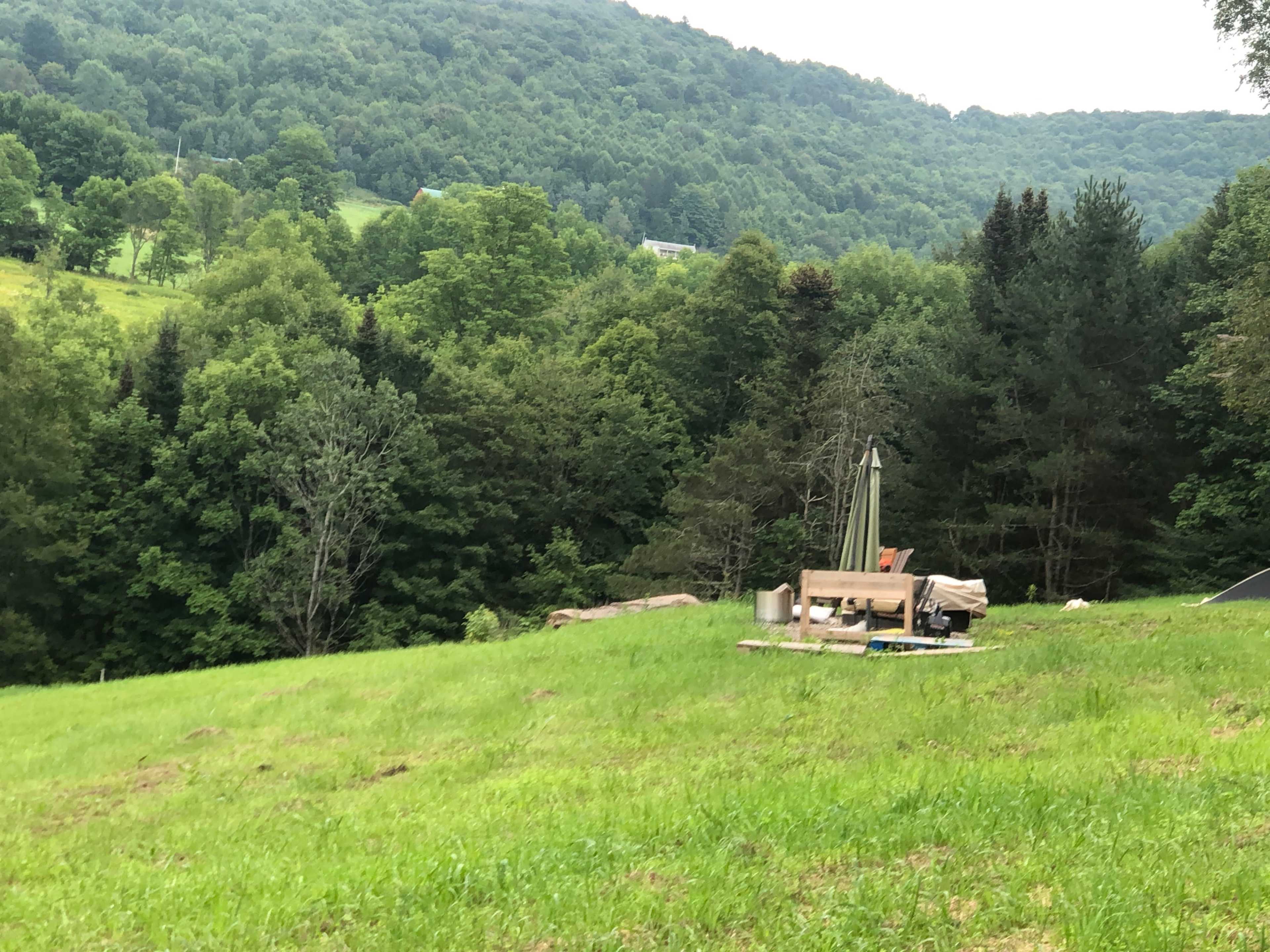 A grassy field with a wooden structure and some camping equipment set against a backdrop of rolling hills and dense trees.