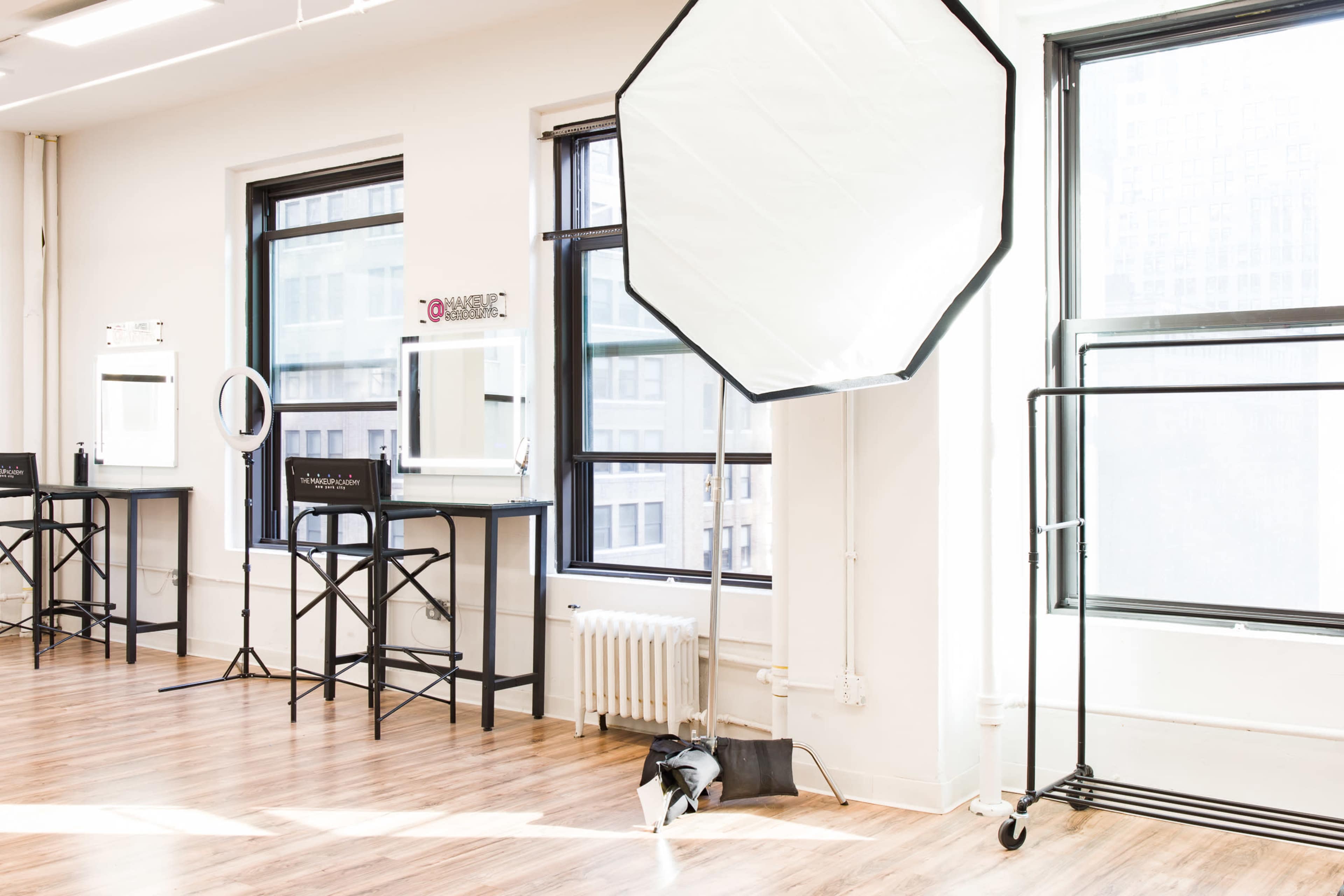 A bright studio space features tall windows, several makeup stations with mirrors, and a large light reflector set up near a radiator.