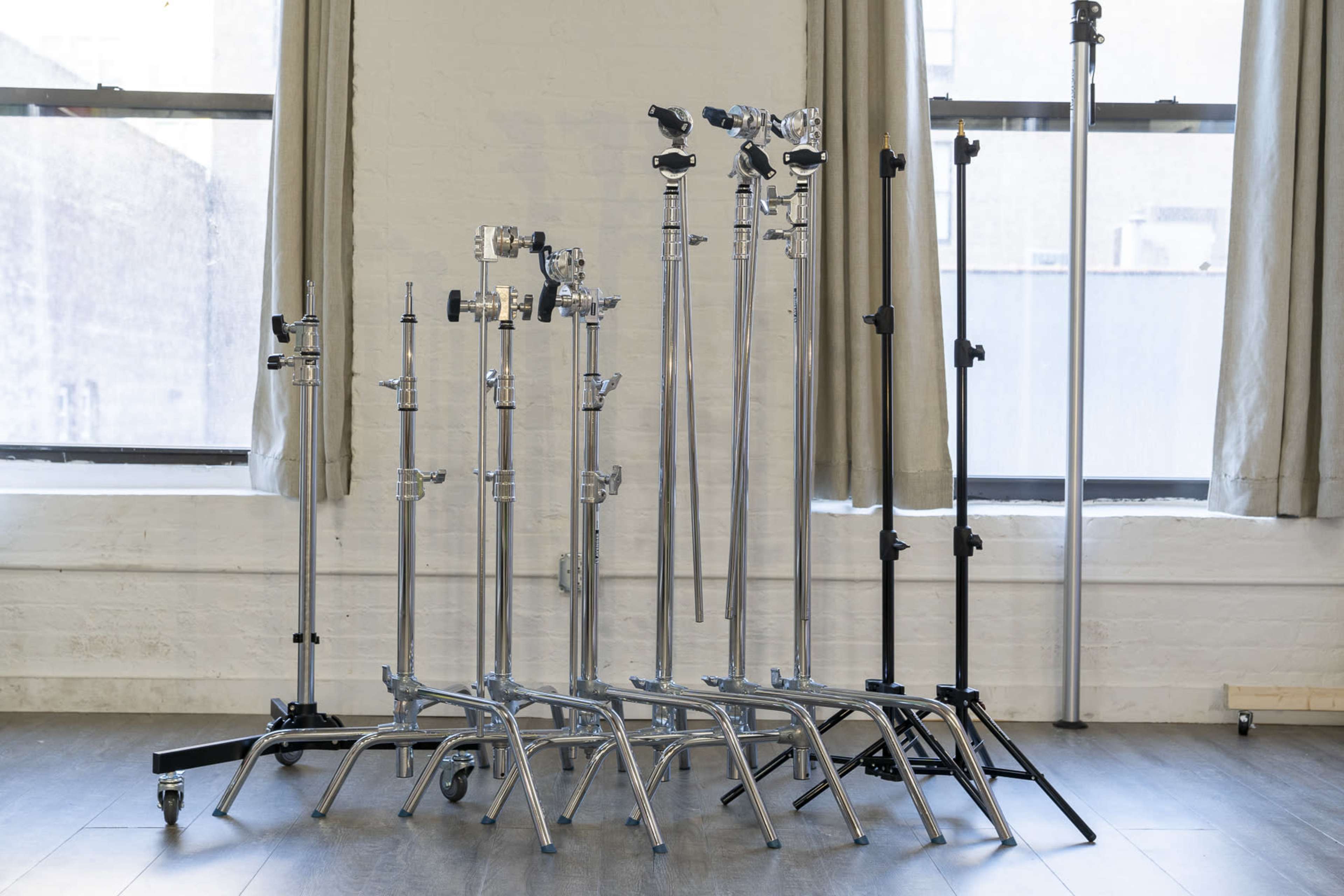 A group of various photography light stands of different heights are positioned near large windows in a studio setting.