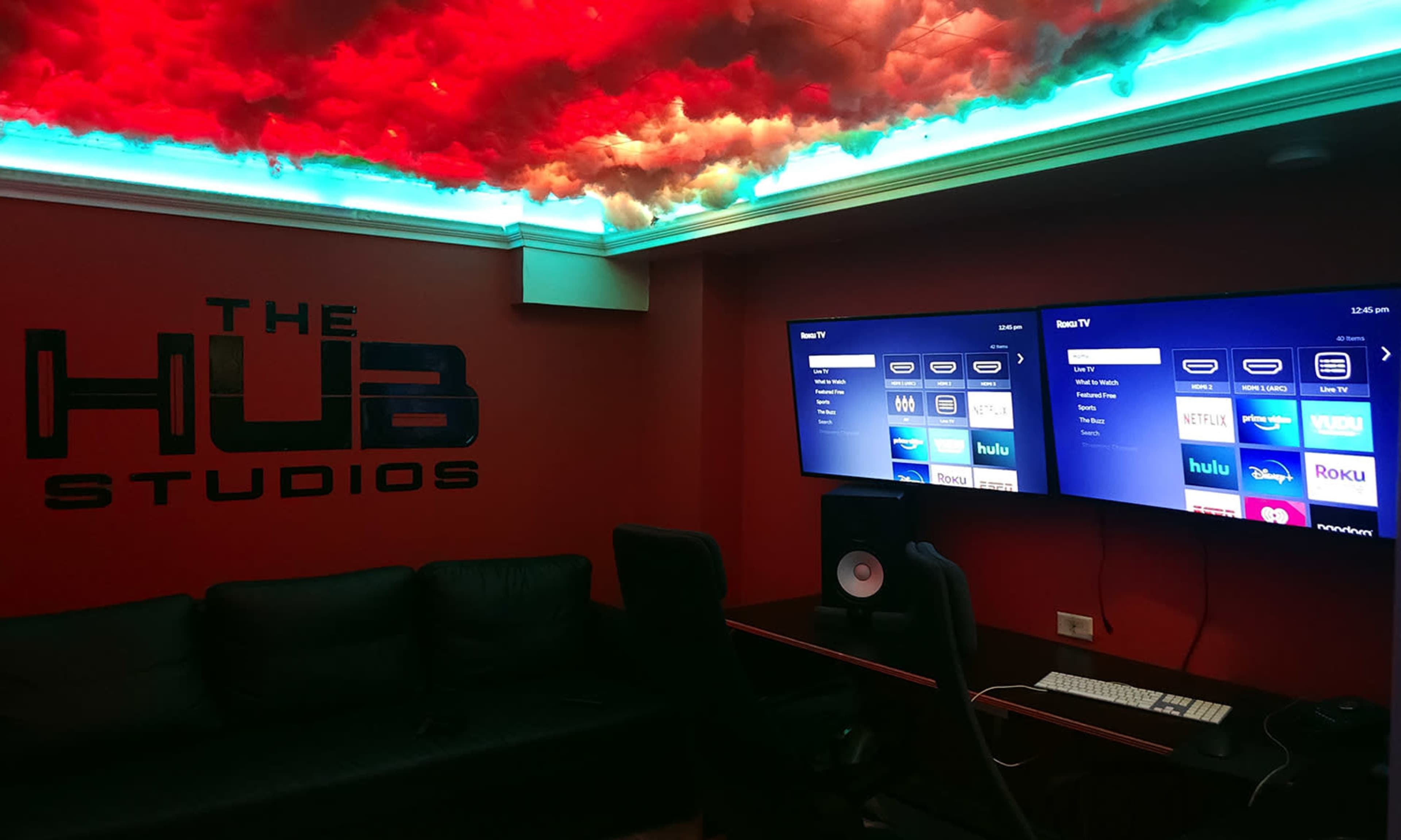 A room labeled "THE HUB STUDIOS," featuring two screens displaying streaming service options, a couch, and a computer setup under a lit ceiling with clouds.