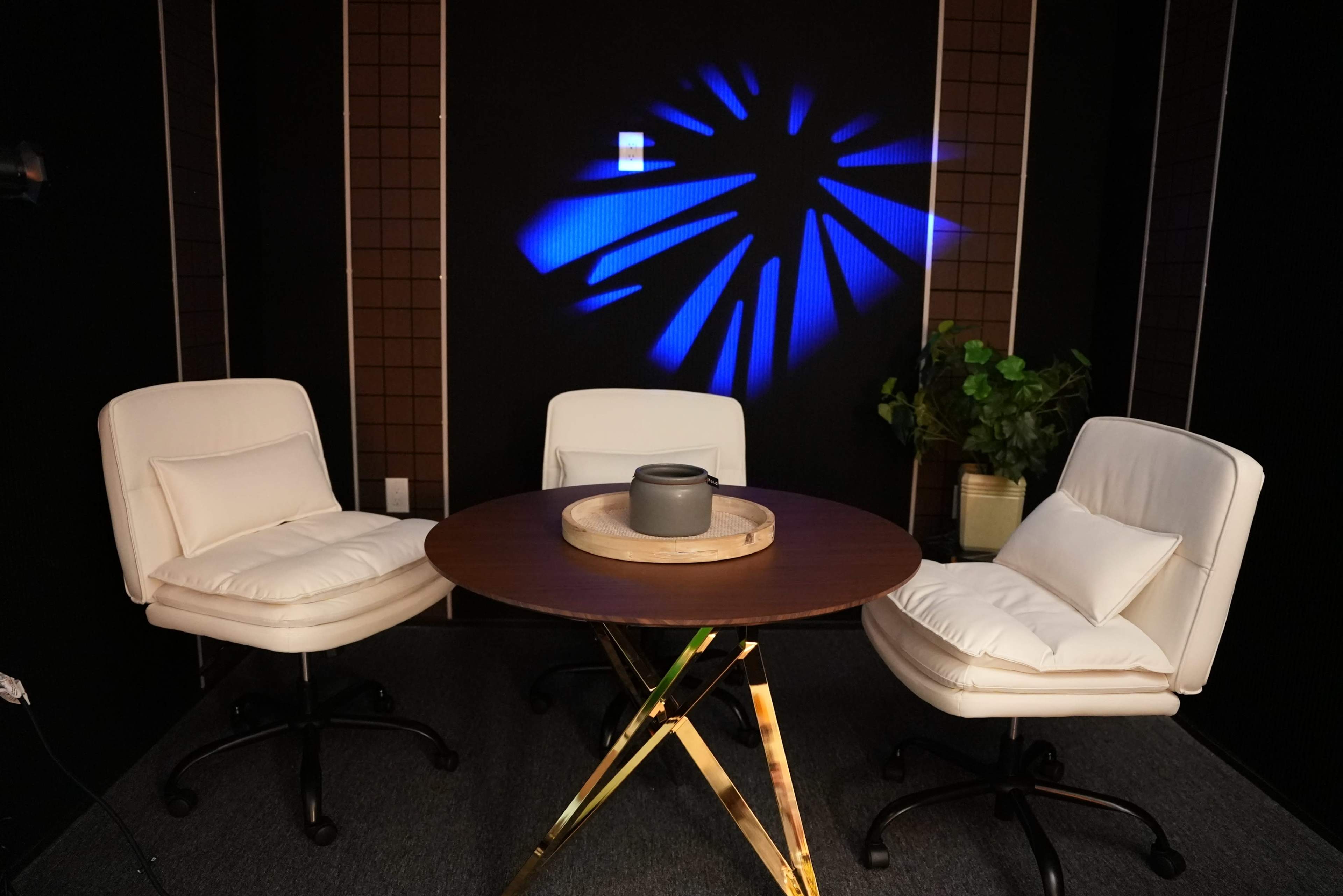 The image shows a small round wooden table surrounded by two white swivel chairs, with a potted plant in the background and blue light casting a pattern on the wall.
