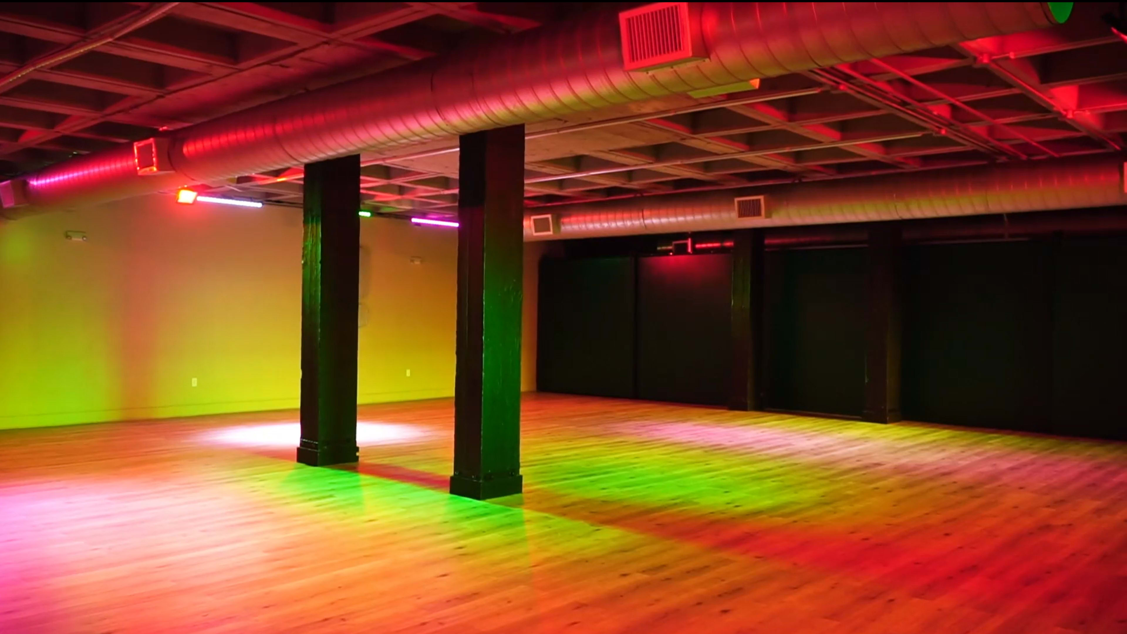 An empty dance studio with wooden floors, illuminated by colorful lights in green and red, and supported by dark wooden pillars.