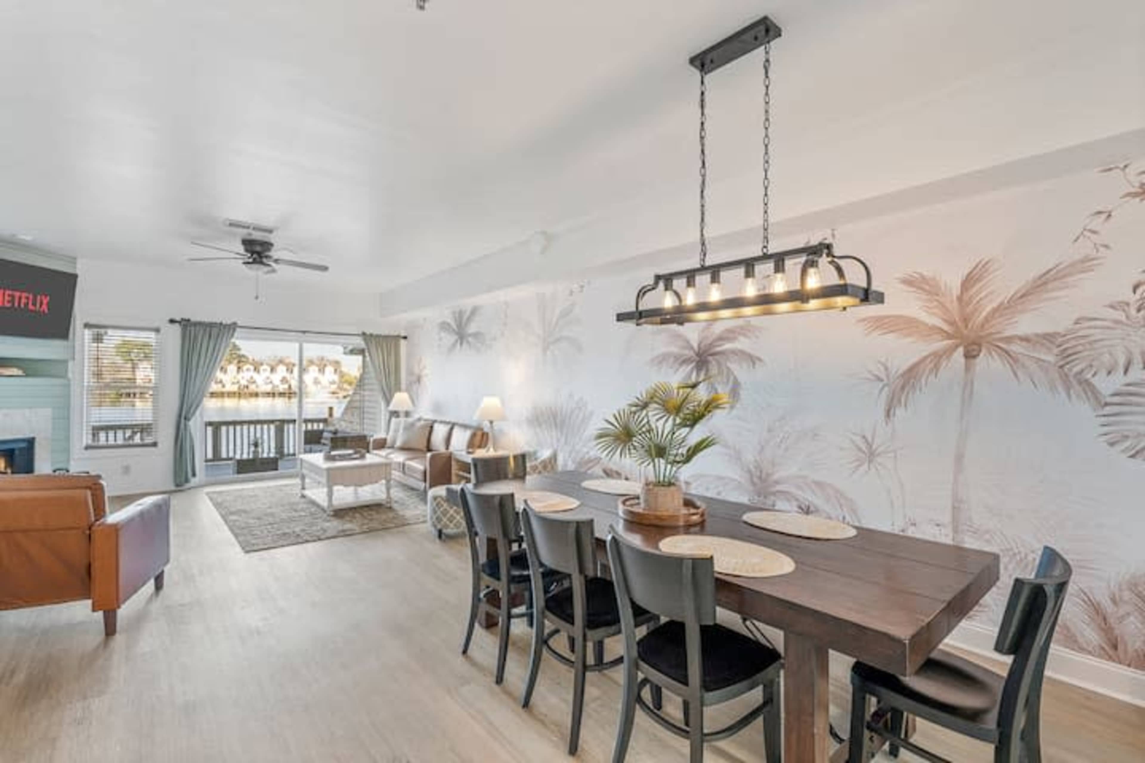 The image shows a spacious living area with a dining table, a sofa, and large windows that overlook a water view, decorated with a palm tree mural.