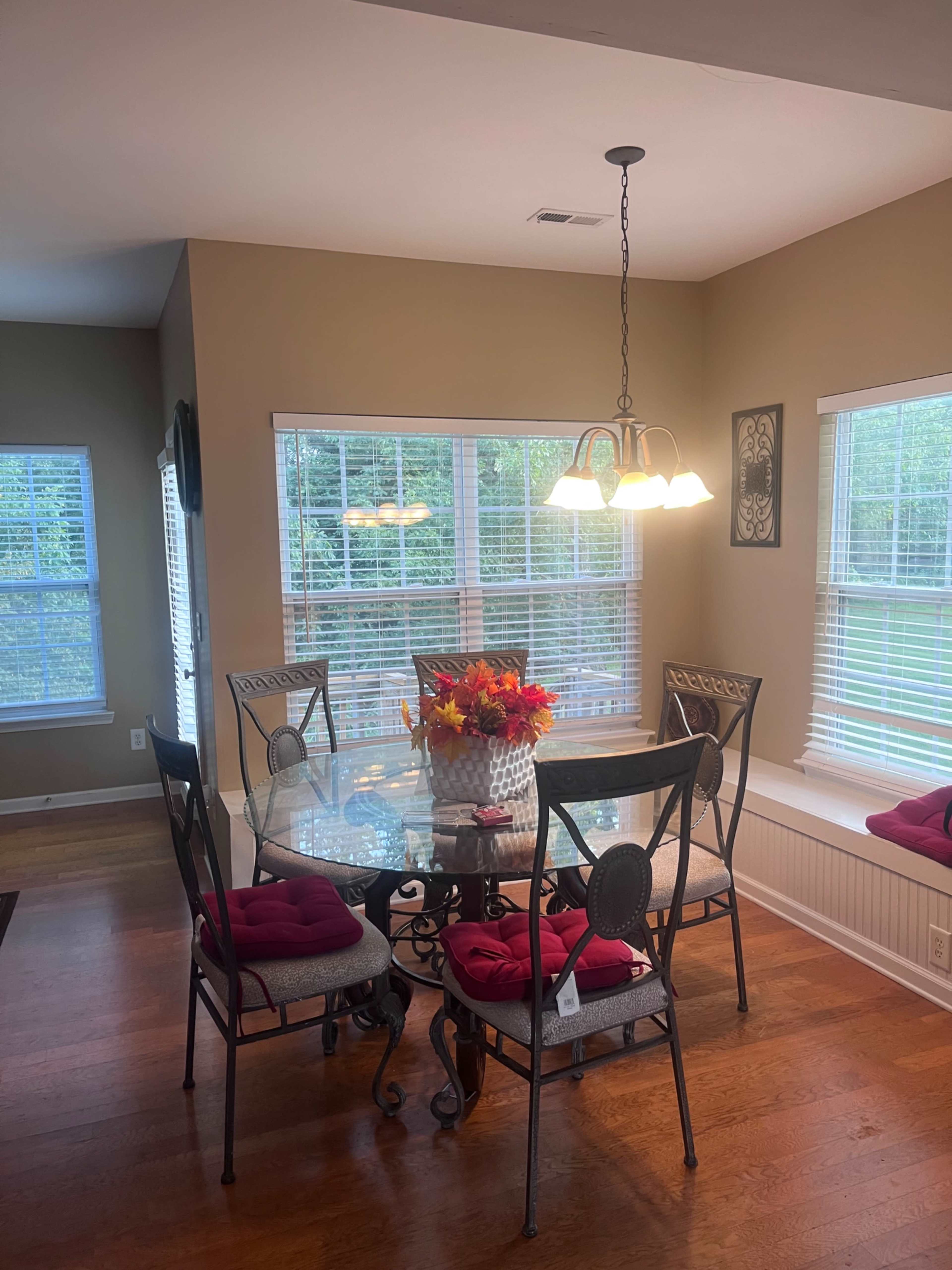 A glass dining table surrounded by six chairs with red cushions is illuminated by a pendant light in a room with multiple windows.
