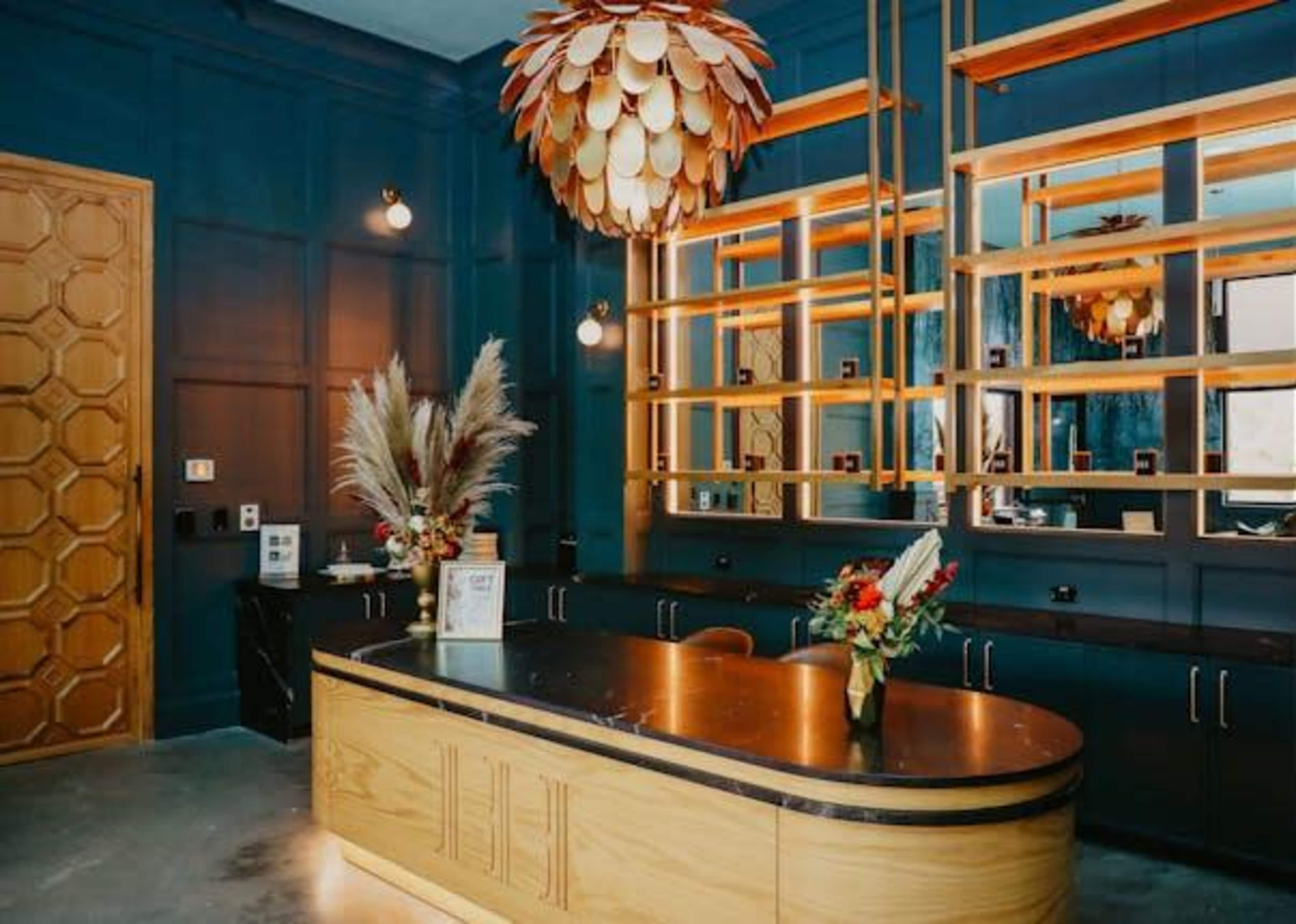 The image shows a contemporary reception area featuring a wooden front desk, large mirrors, a decorative pendant light, and potted plants.