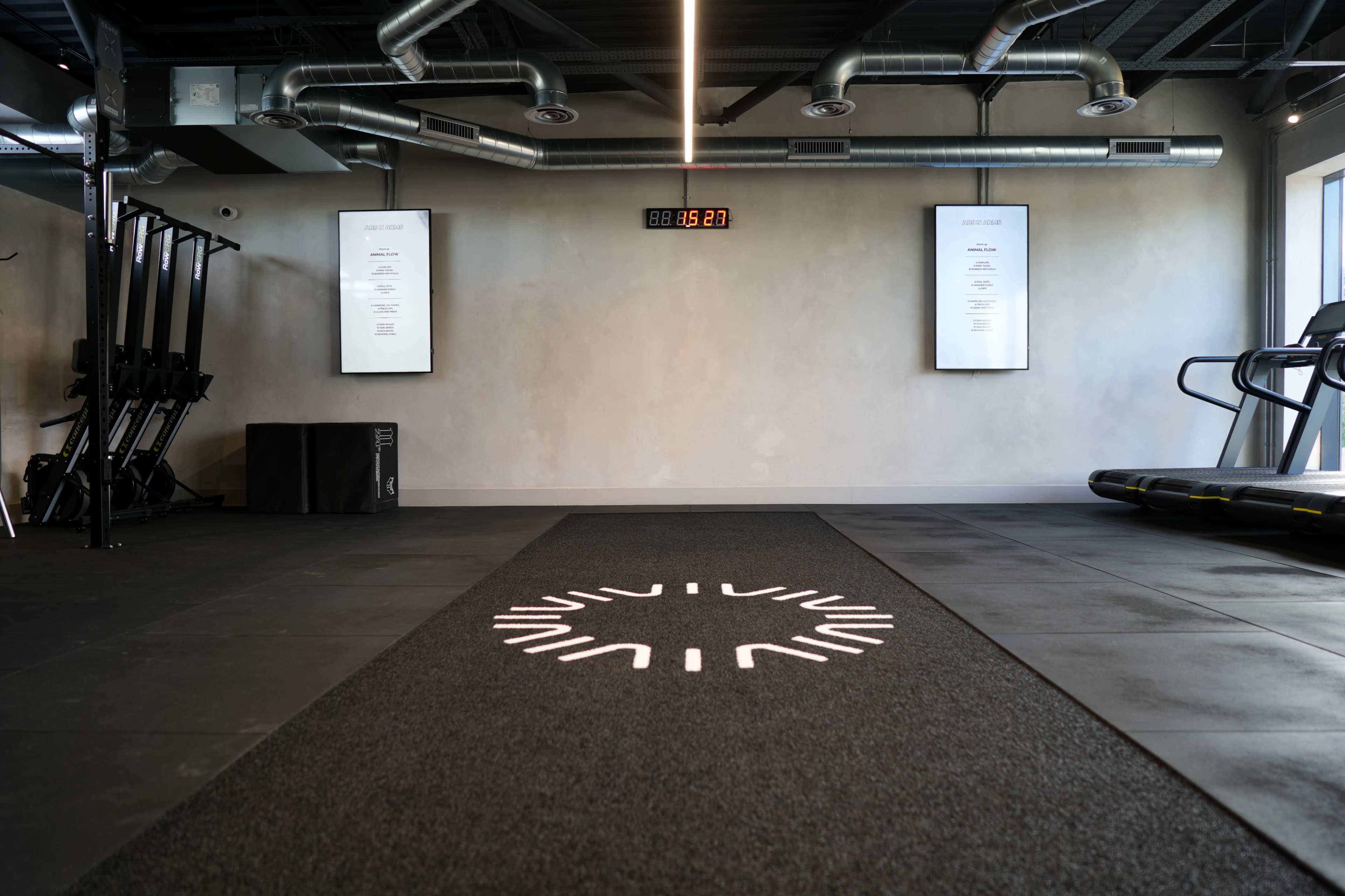 The image shows a modern gym interior with a black workout mat, fitness equipment, and digital displays on the walls.