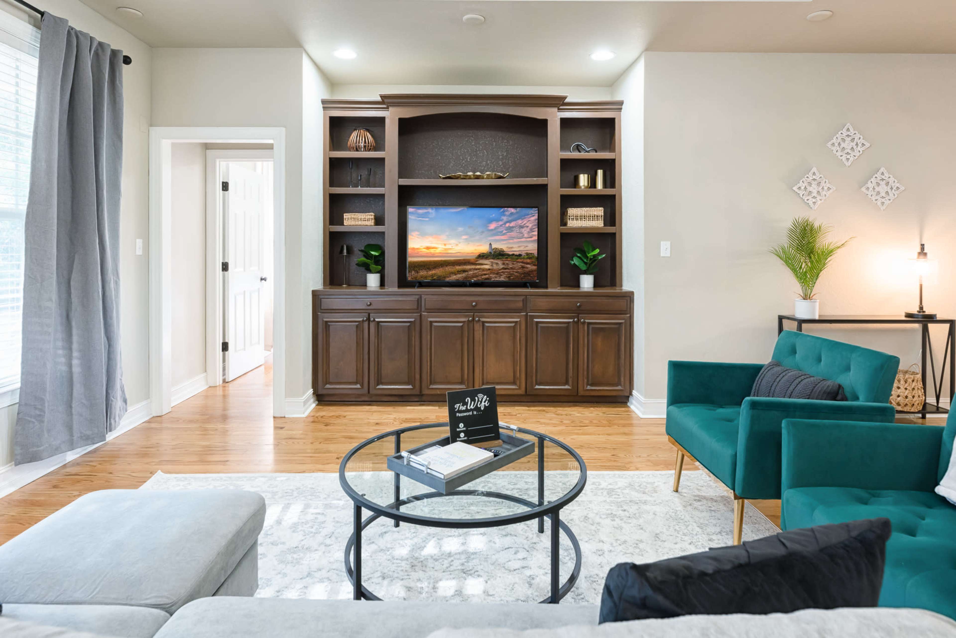 A modern living room features a wooden entertainment center with a large TV displaying a scenic landscape, surrounded by a light gray sectional sofa, green chairs, and a circular coffee table on a textured rug.