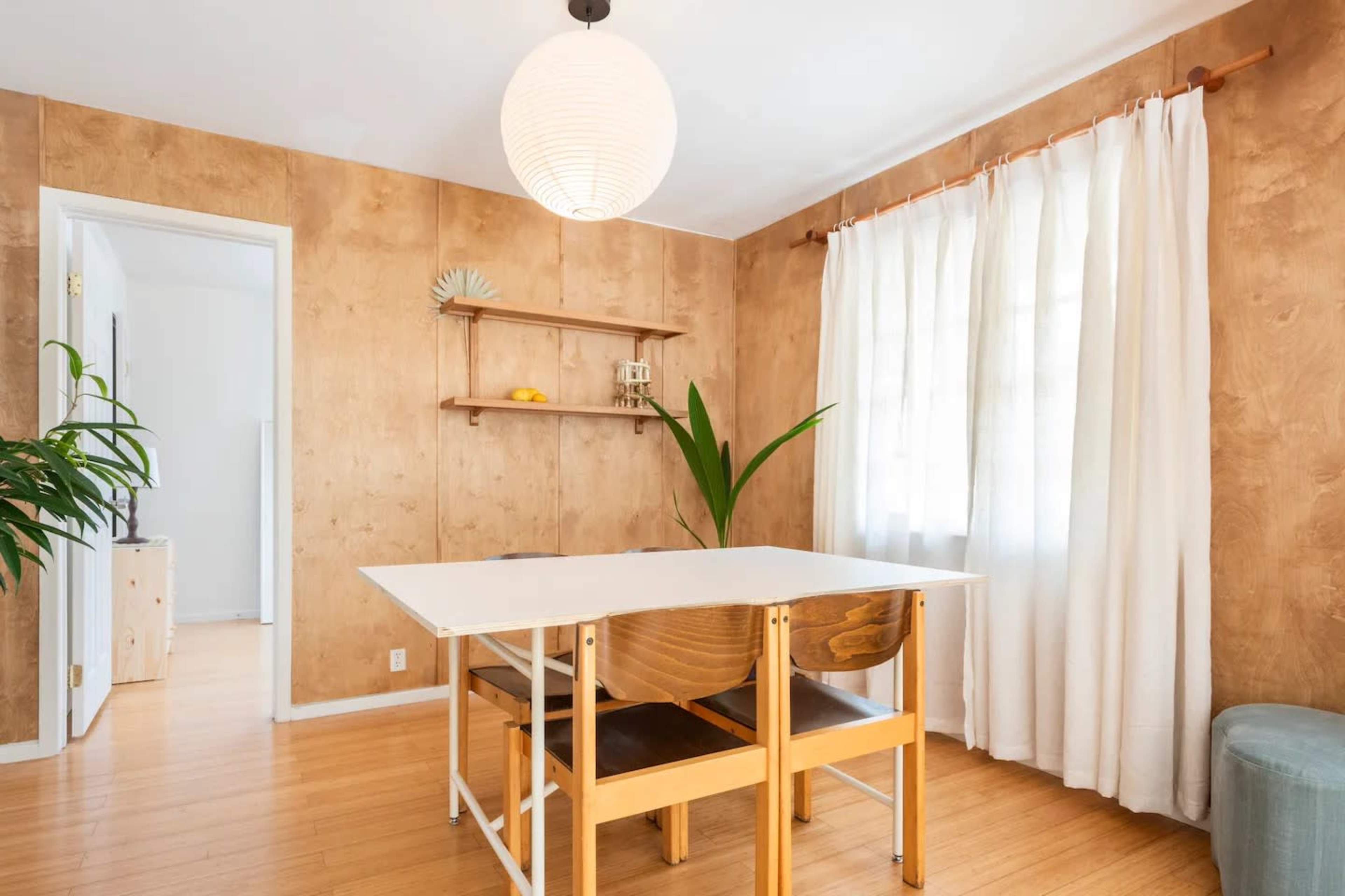 A dining area with a white table and wooden chairs, surrounded by light-colored walls, a window with sheer curtains, and a shelf displaying decor.