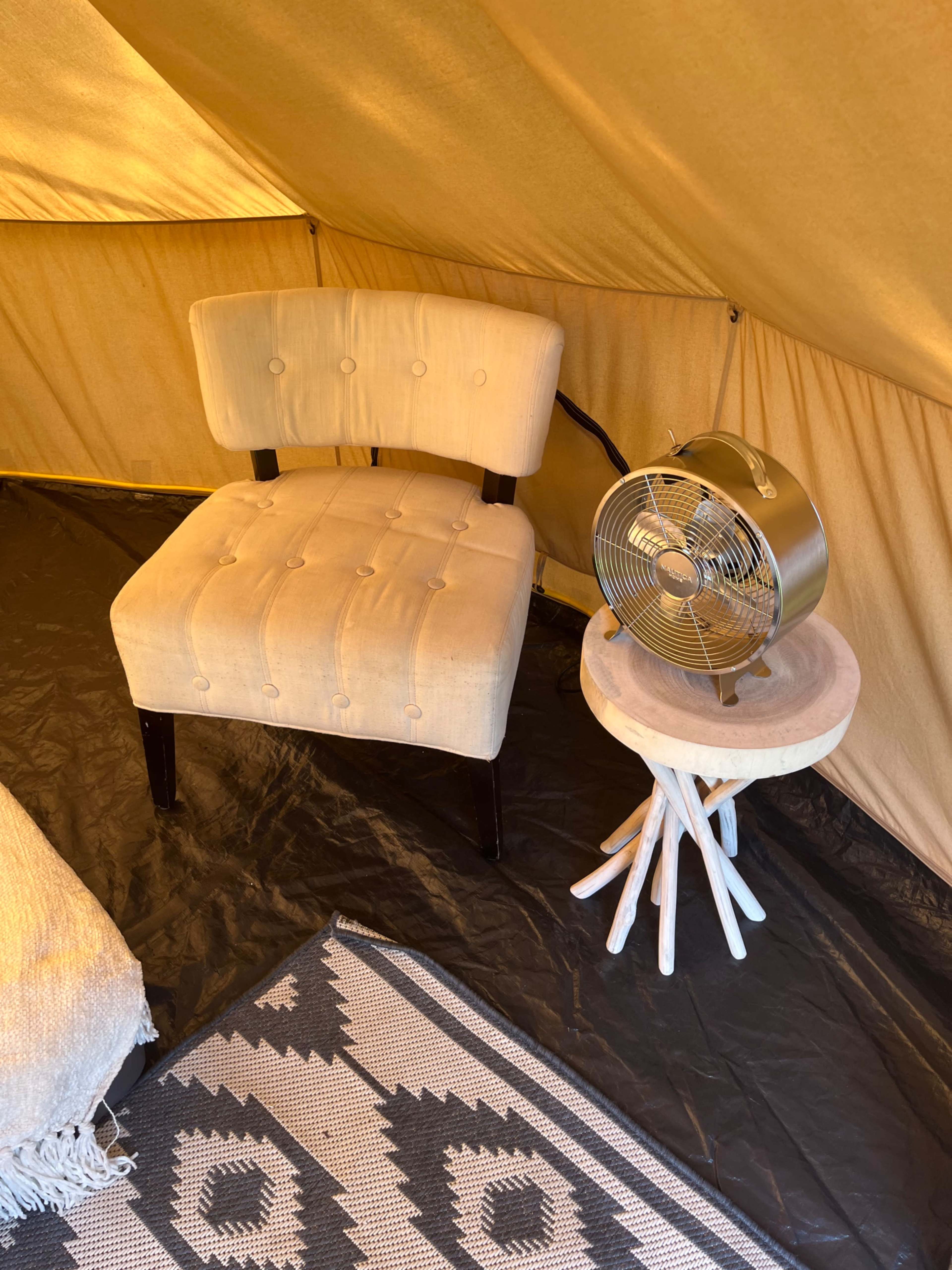An interior of a tent featuring a beige upholstered chair, a small wooden side table with a fan, and a patterned rug on a black floor.