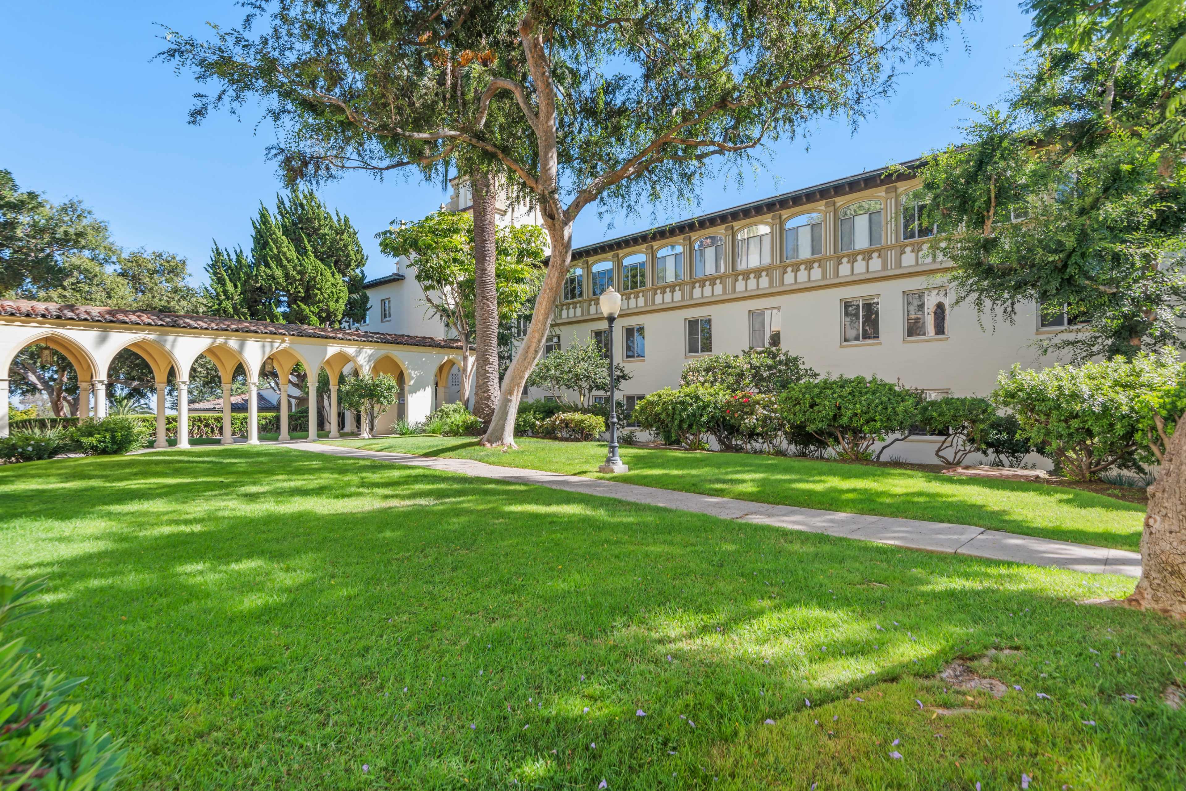 The image shows a manicured lawn with a walkway leading to a building featuring arches and multiple windows, surrounded by trees and bushes.