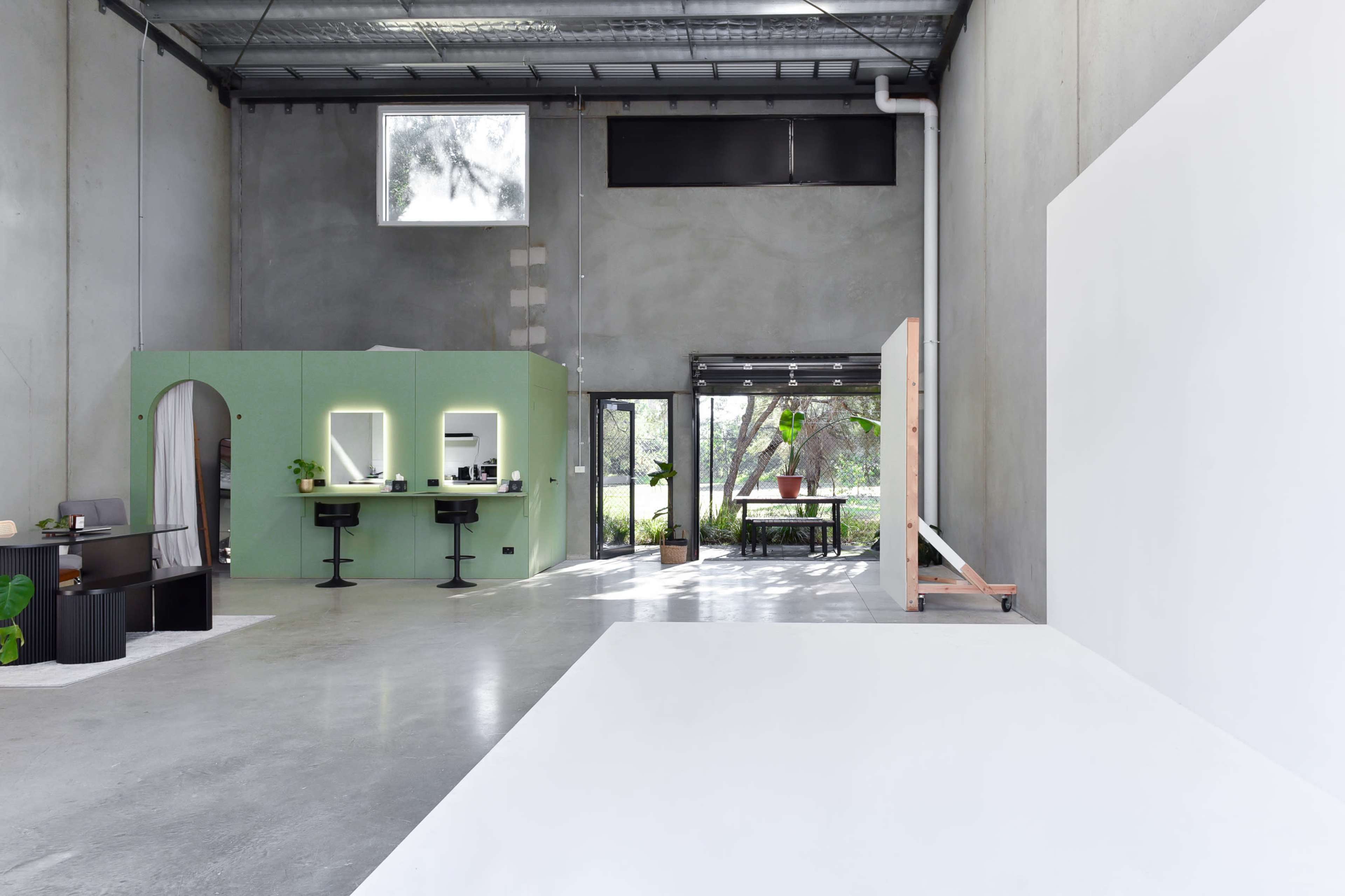 A spacious, industrial-style interior with a green partition housing mirrors and black stools, leading to an area with a large white backdrop and natural light coming through large windows.