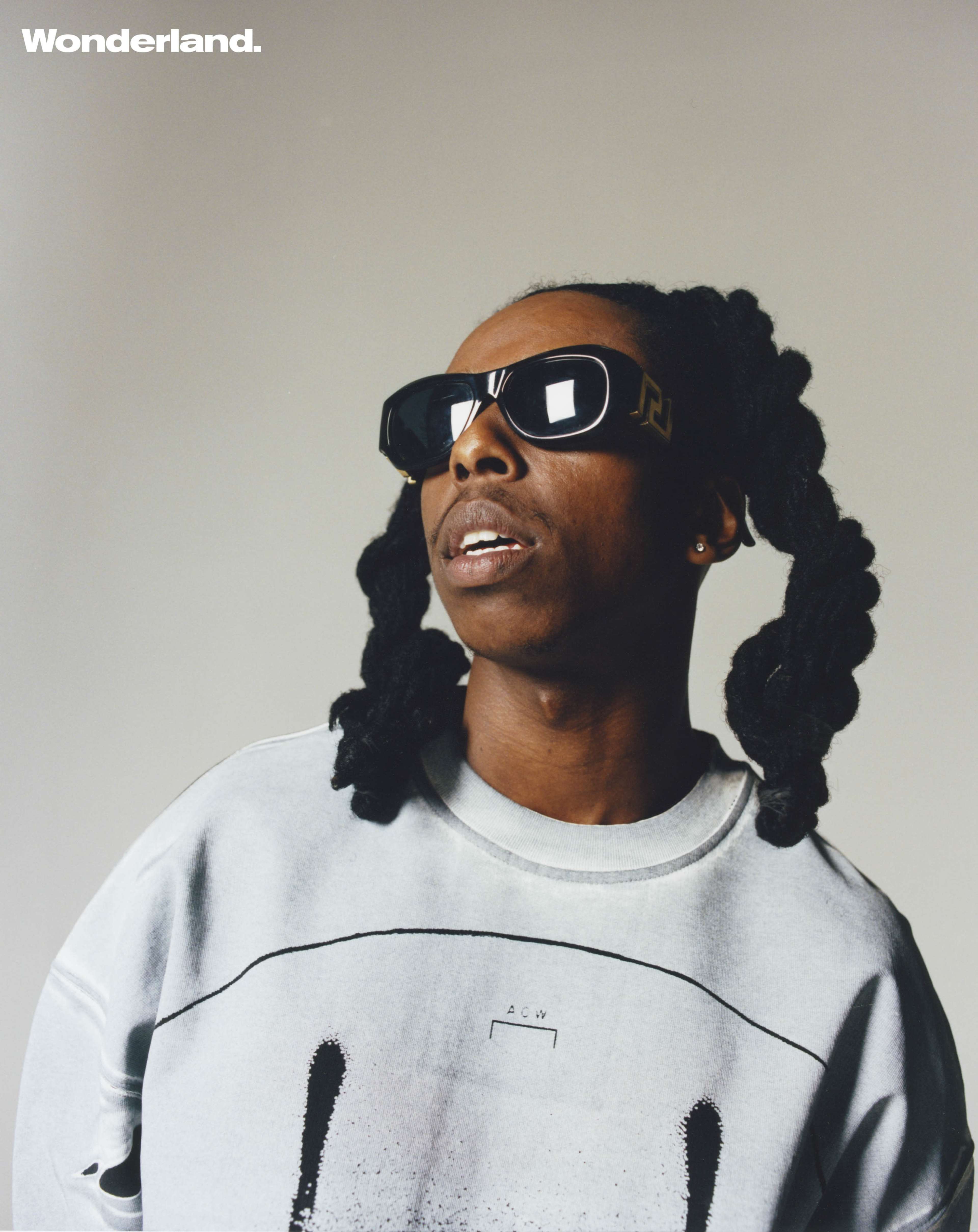 A person with long, styled hair wears sunglasses and a sweatshirt while gazing upward against a plain background.