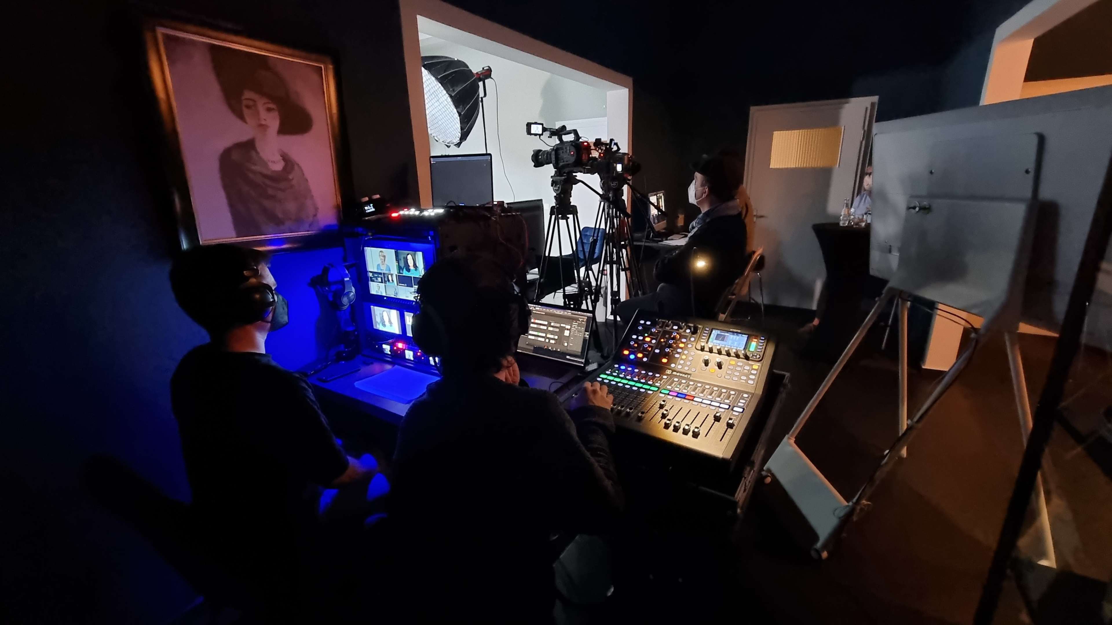 A film or video production setup, featuring multiple cameras and production equipment in a dimly lit room with a portrait hanging on the wall.