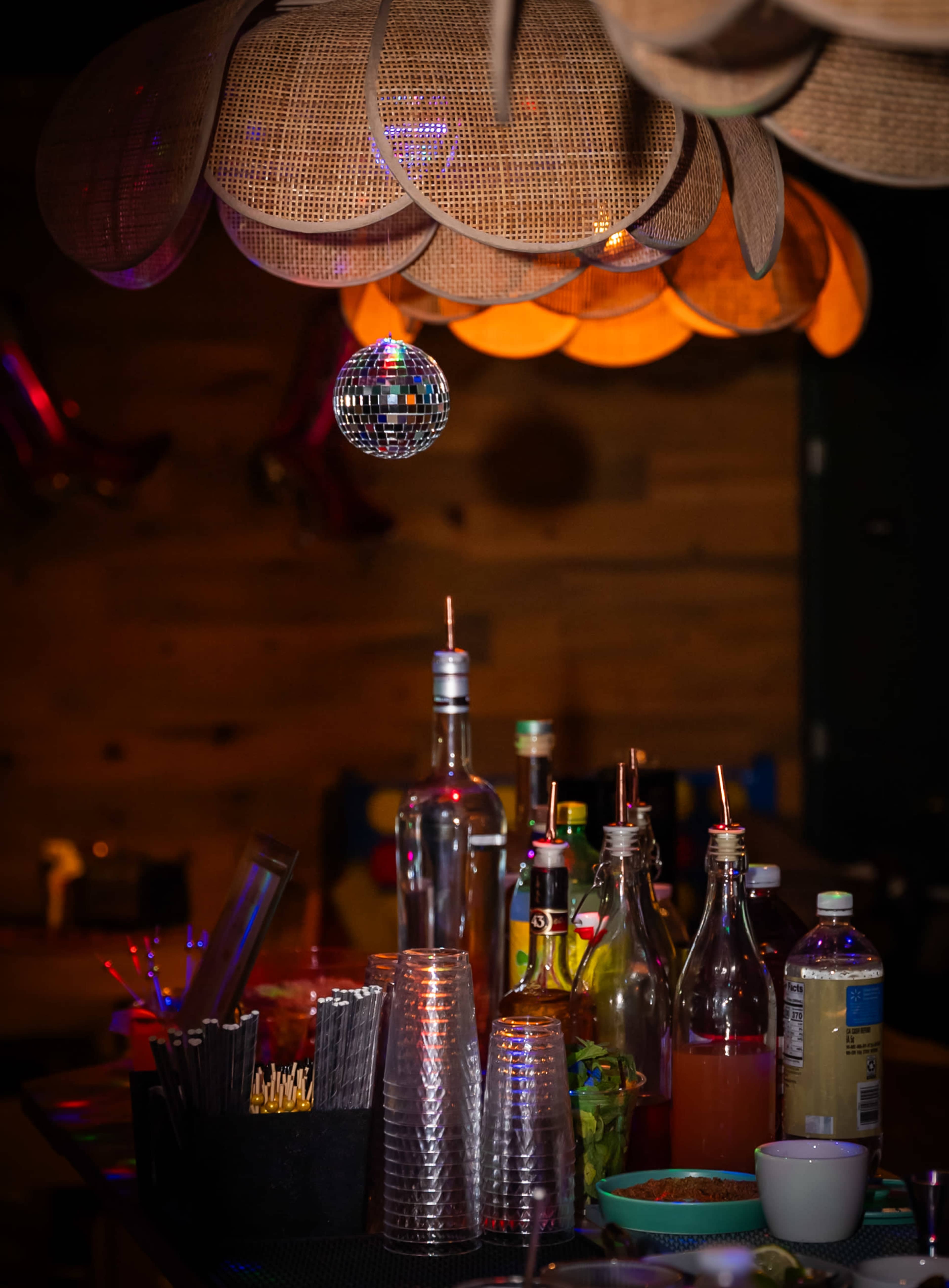 A bar counter with various bottles and glasses under a decorative light fixture, featuring a disco ball hanging above.