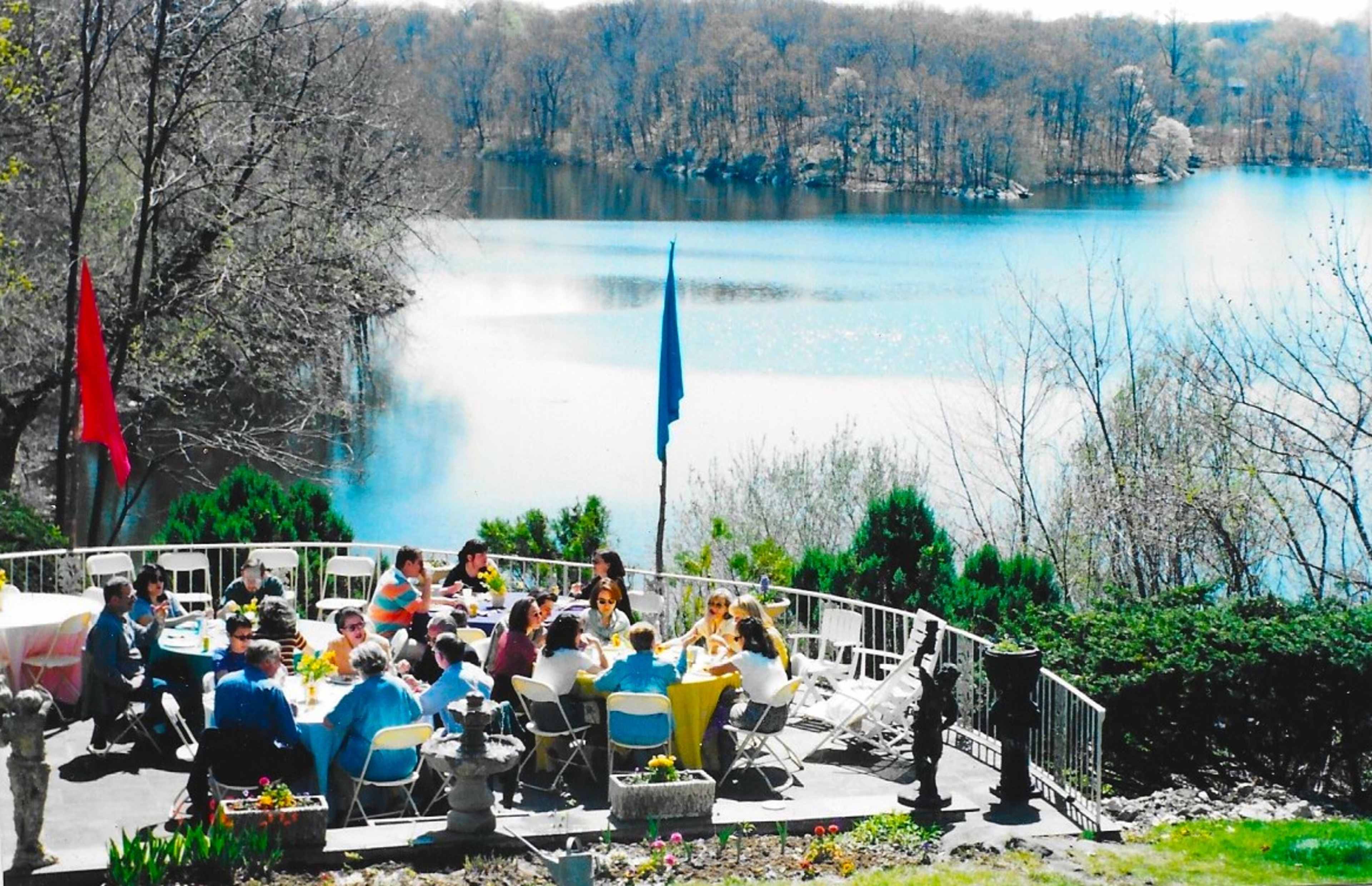 A large group of people sits at tables on a patio overlooking a calm lake surrounded by trees.