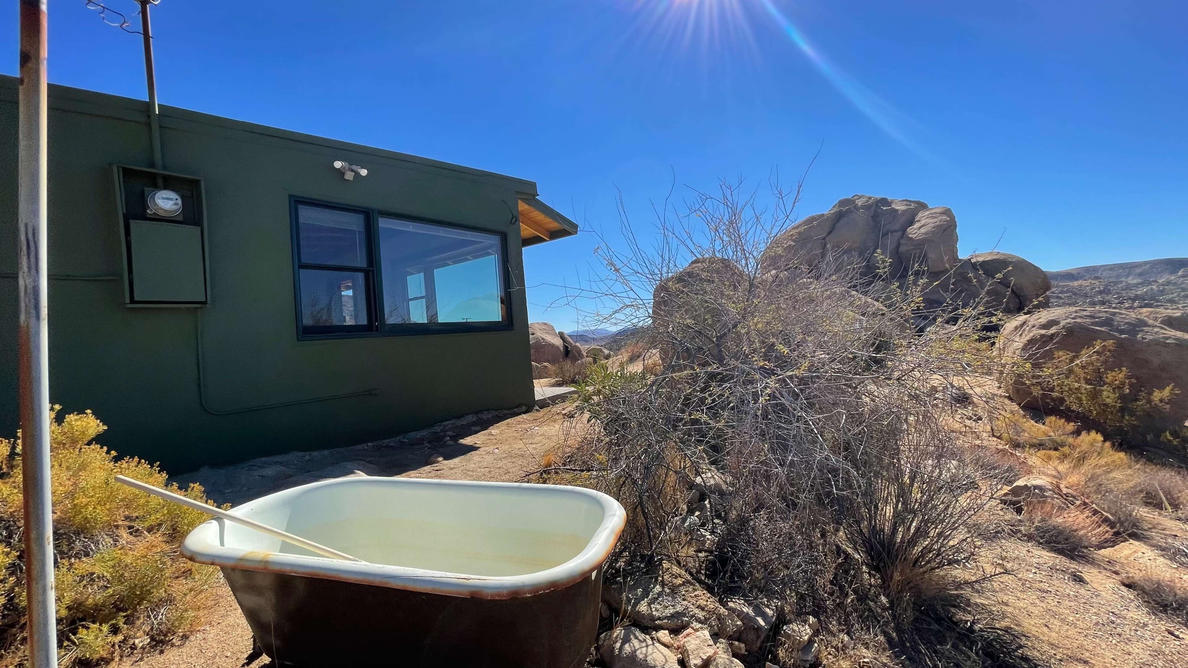 A vintage metal bathtub sits on a rocky pathway beside a green building and boulders under a clear blue sky.