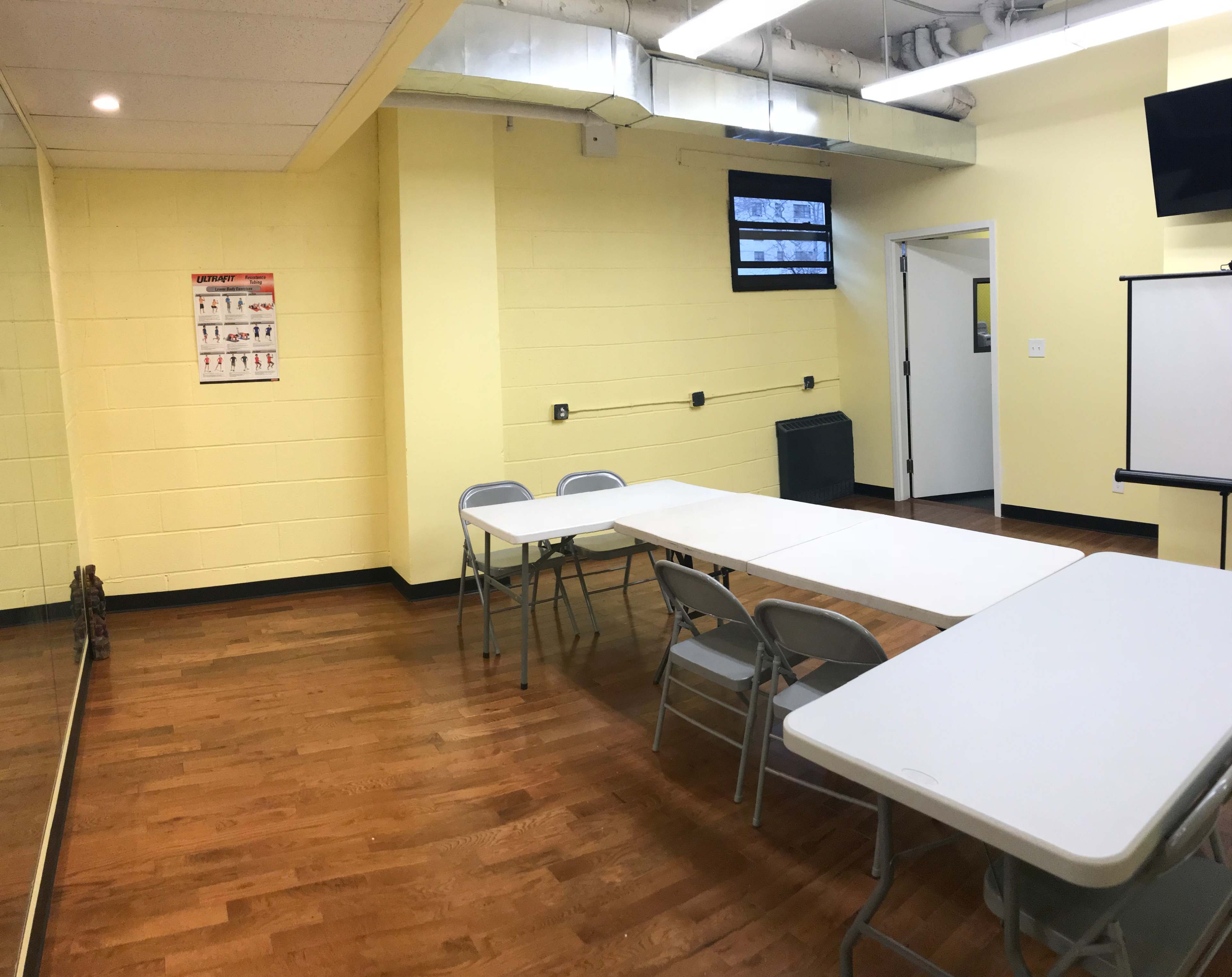 A small classroom or meeting room with yellow walls, a few folding tables, chairs, and a mirror on one side.