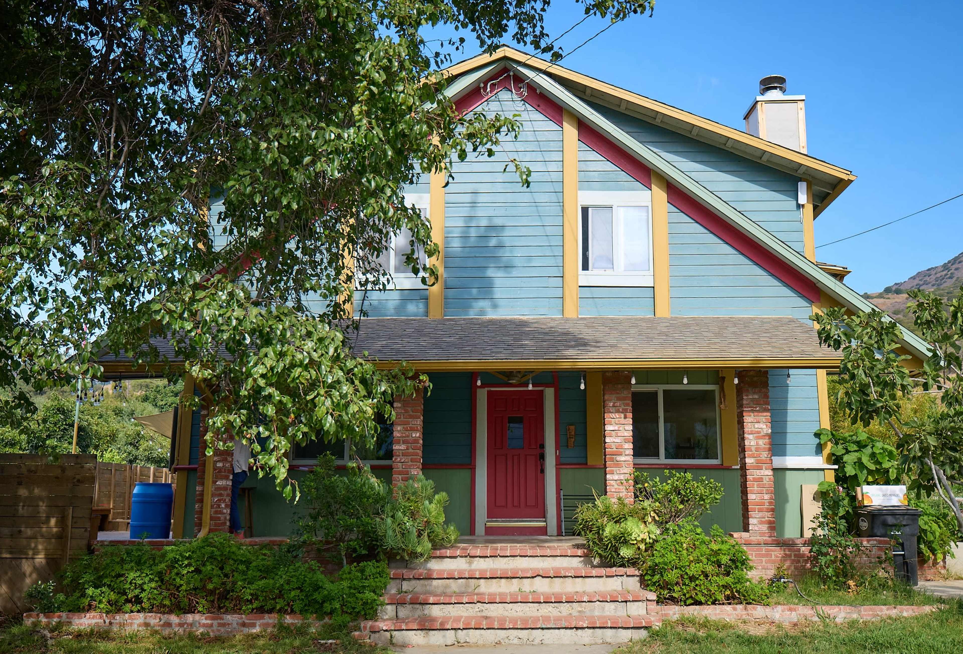 The image shows a multi-colored two-story house with a red door and stairs leading up to it, surrounded by greenery.