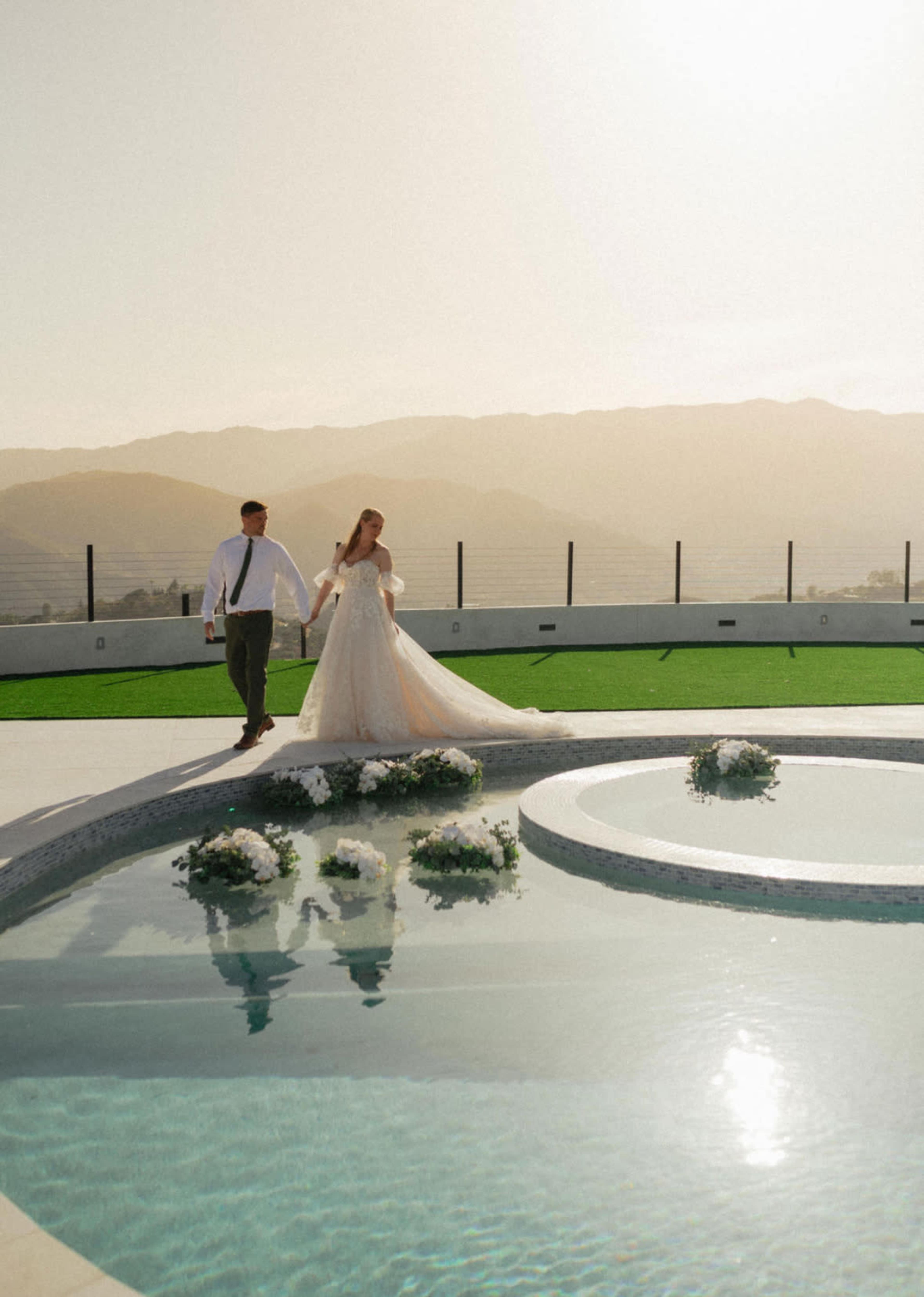 A couple walks hand in hand beside a circular pool on a rooftop with mountains in the background.