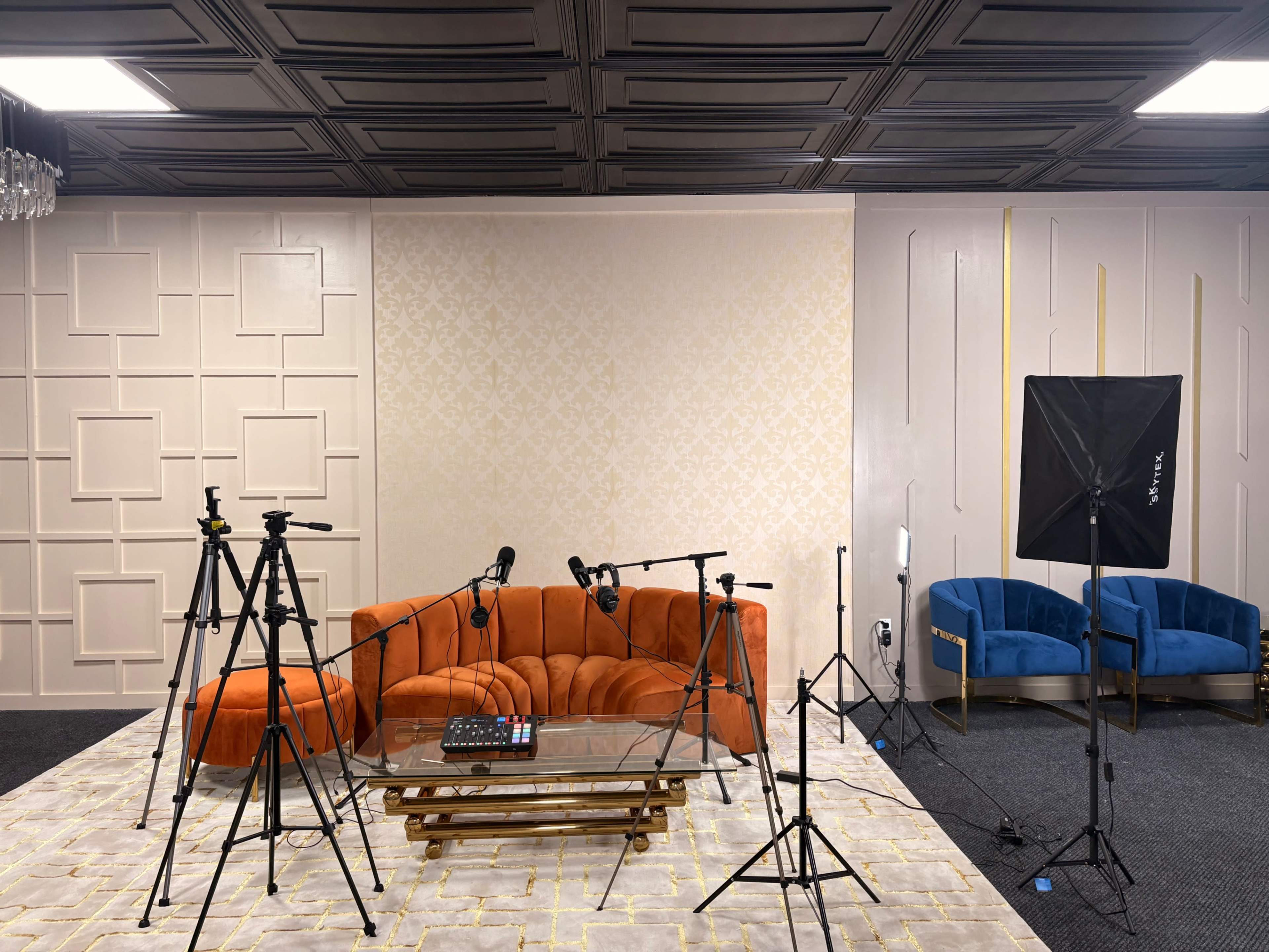 The image shows a modern studio setup with an orange couch, multiple tripods with cameras and microphones, and blue chairs against a decorative wall backdrop.