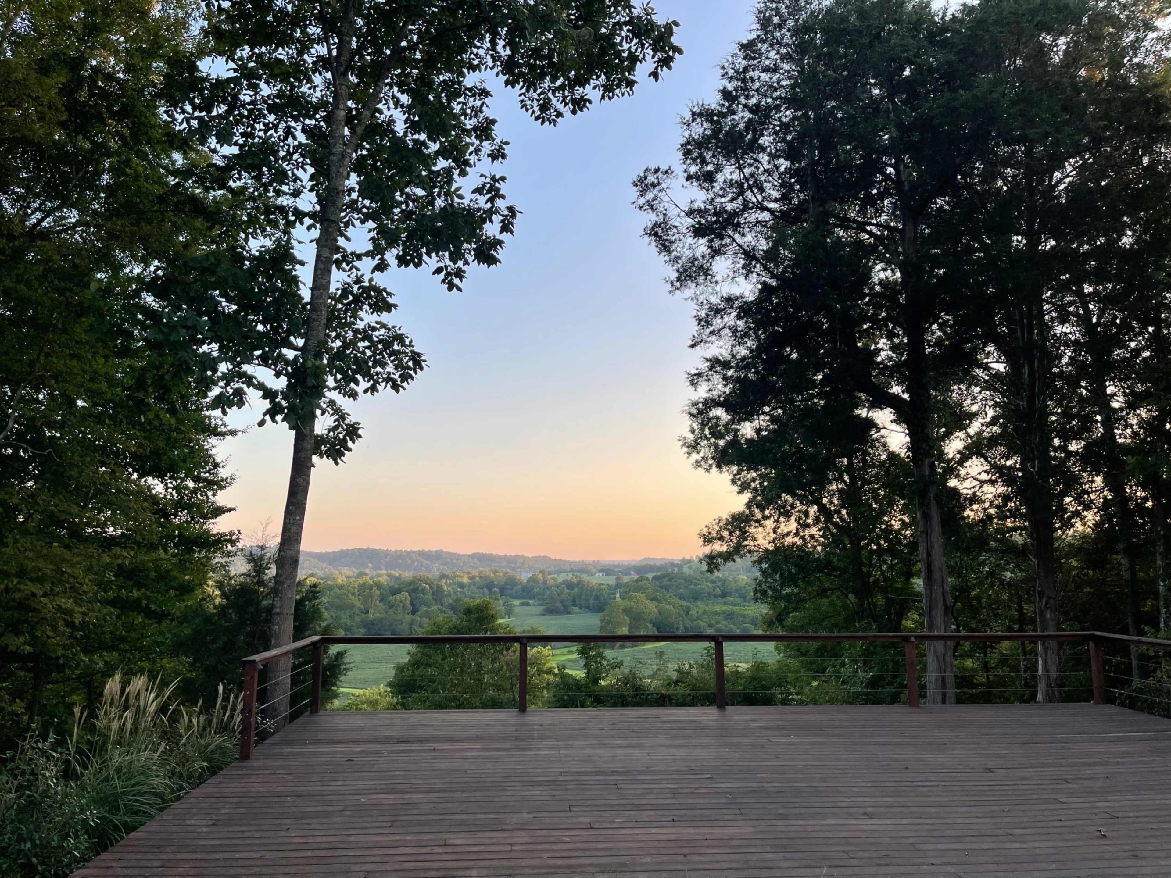 A wooden deck overlooks a green valley surrounded by trees against a serene sky at dusk.