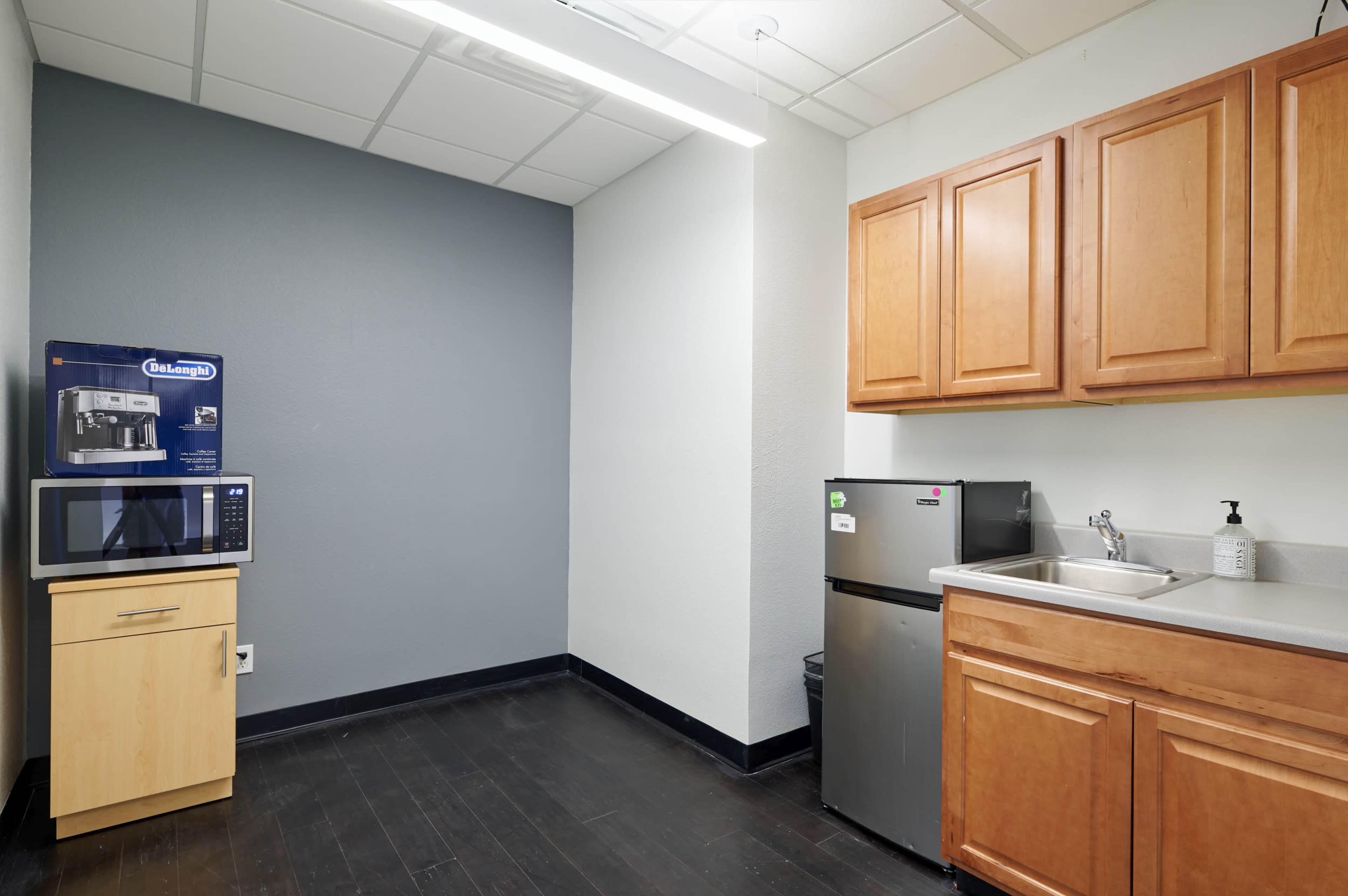 The image shows a small kitchenette with wooden cabinets, a microwave, a refrigerator, and a sink against a gray wall.