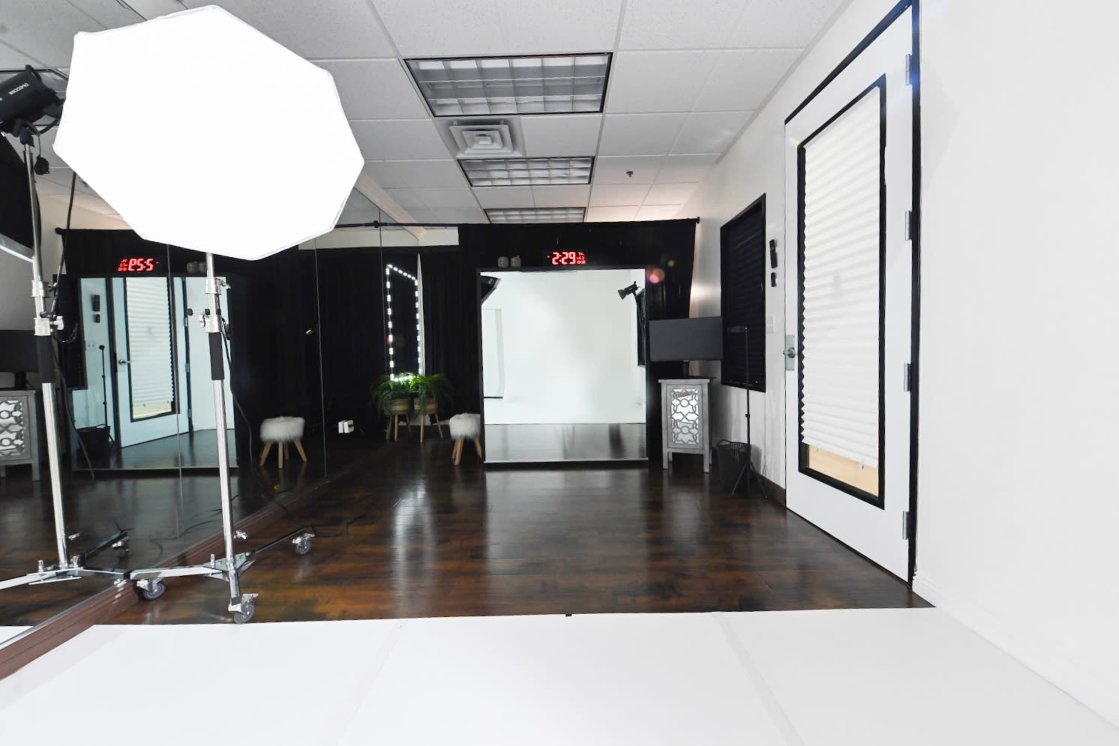 The image shows a spacious photography studio with a large mirror, a softbox light, and a closed door, featuring a white floor and dark wooden walls.