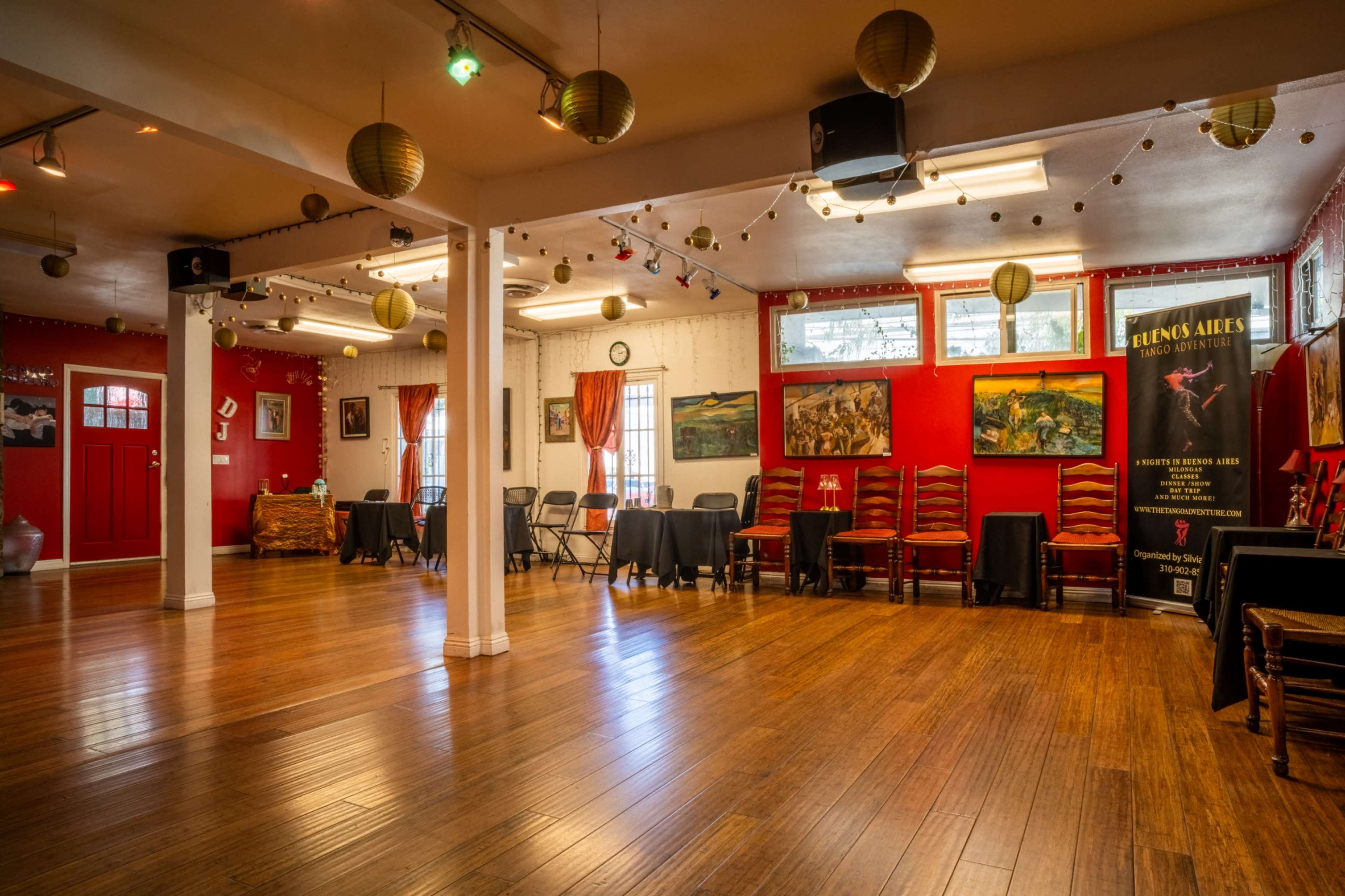 The image shows a spacious, well-lit room with wooden floors, adorned with chairs and tables, and decorated with art pieces and lanterns hanging from the ceiling.